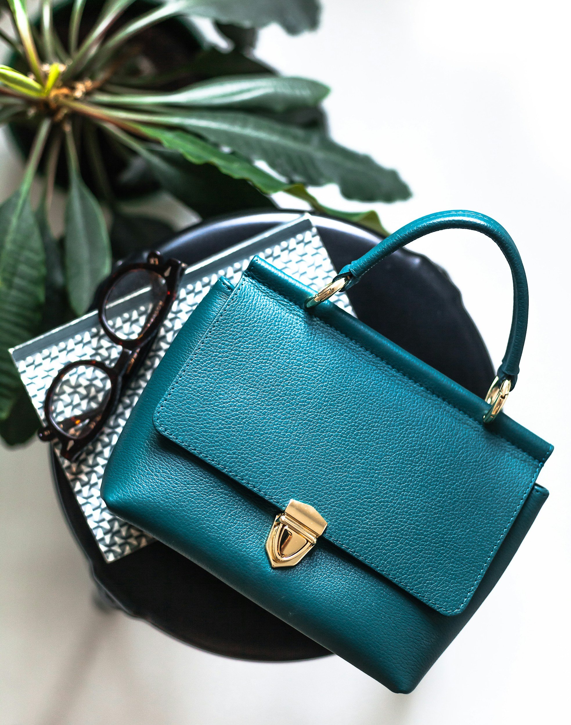 A teal handbag with gold hardware, a pair of tortoiseshell glasses, and a silver patterned notebook resting on a black round surface, with green leaves in the background.