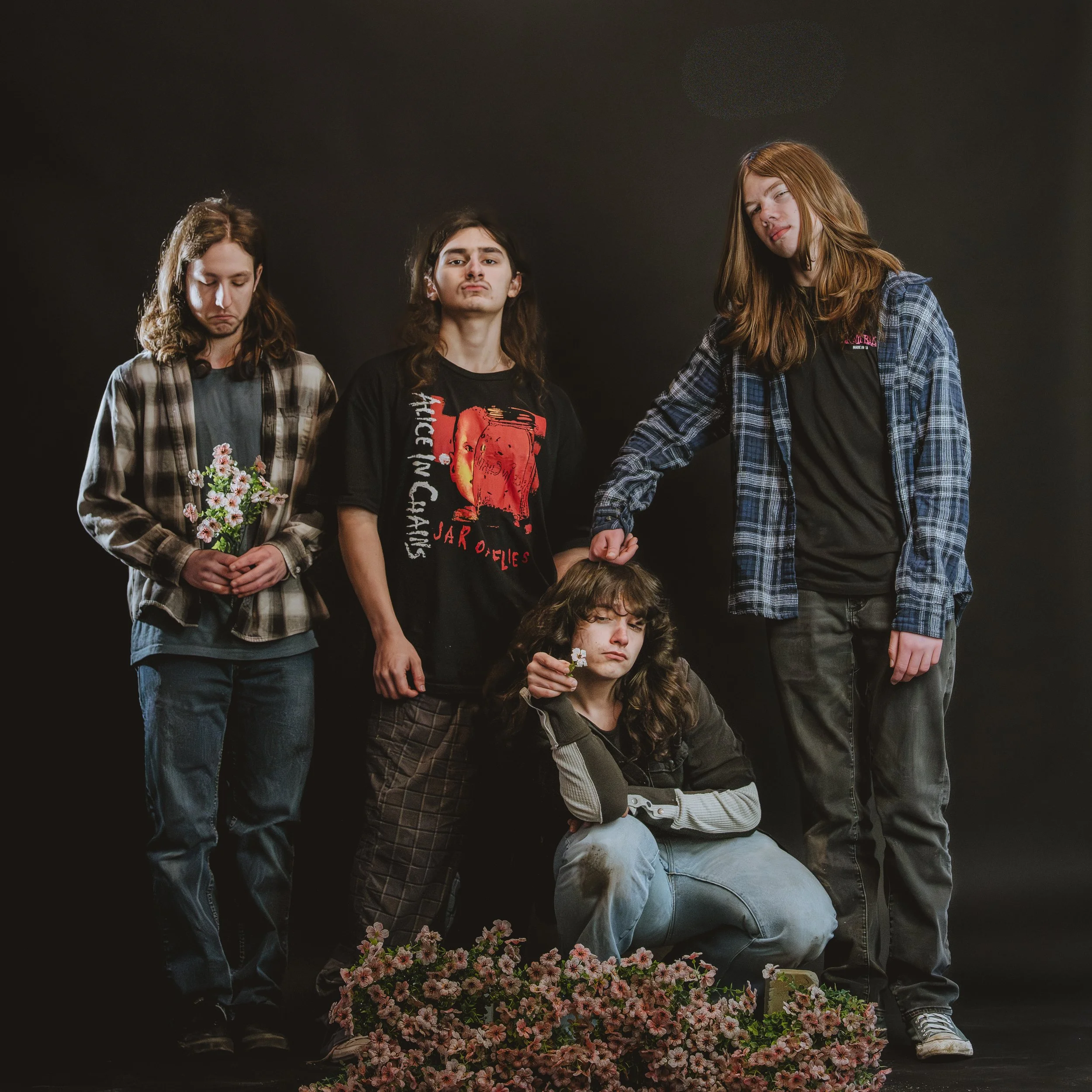 Four young people with long hair standing in front of a dark background, some holding flowers and making various facial expressions.