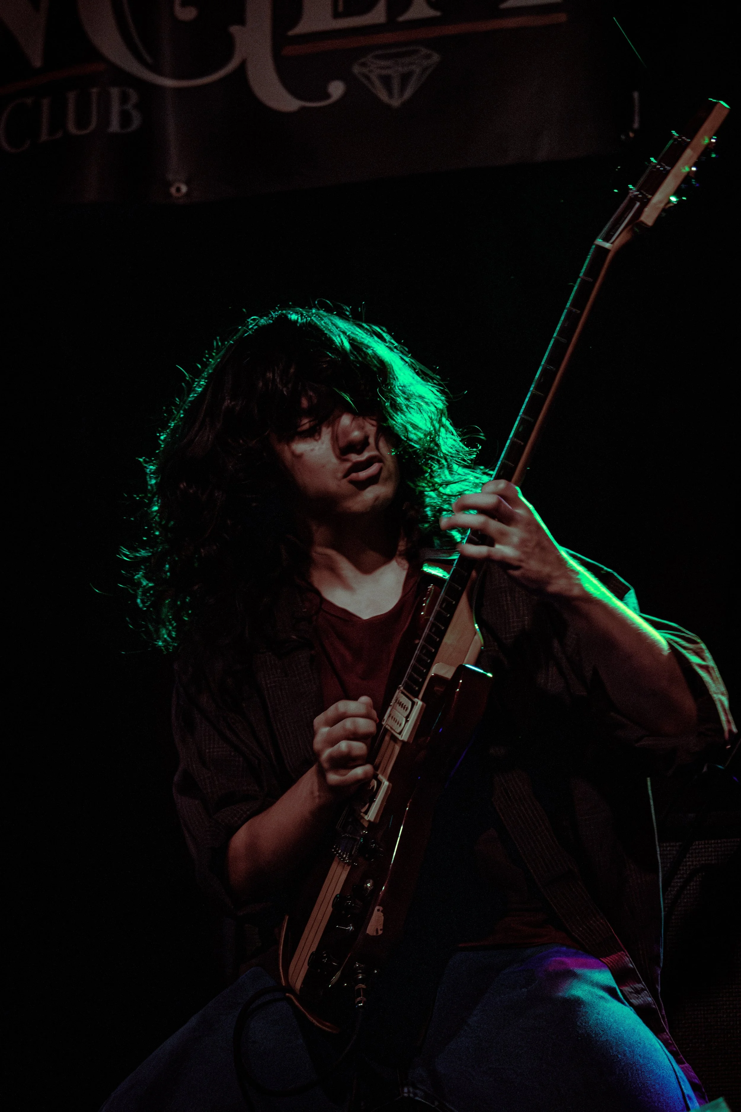 Musician playing an electric guitar on stage with green lighting highlighting curly hair and face, dark background, and a sign partially visible in the upper left corner.