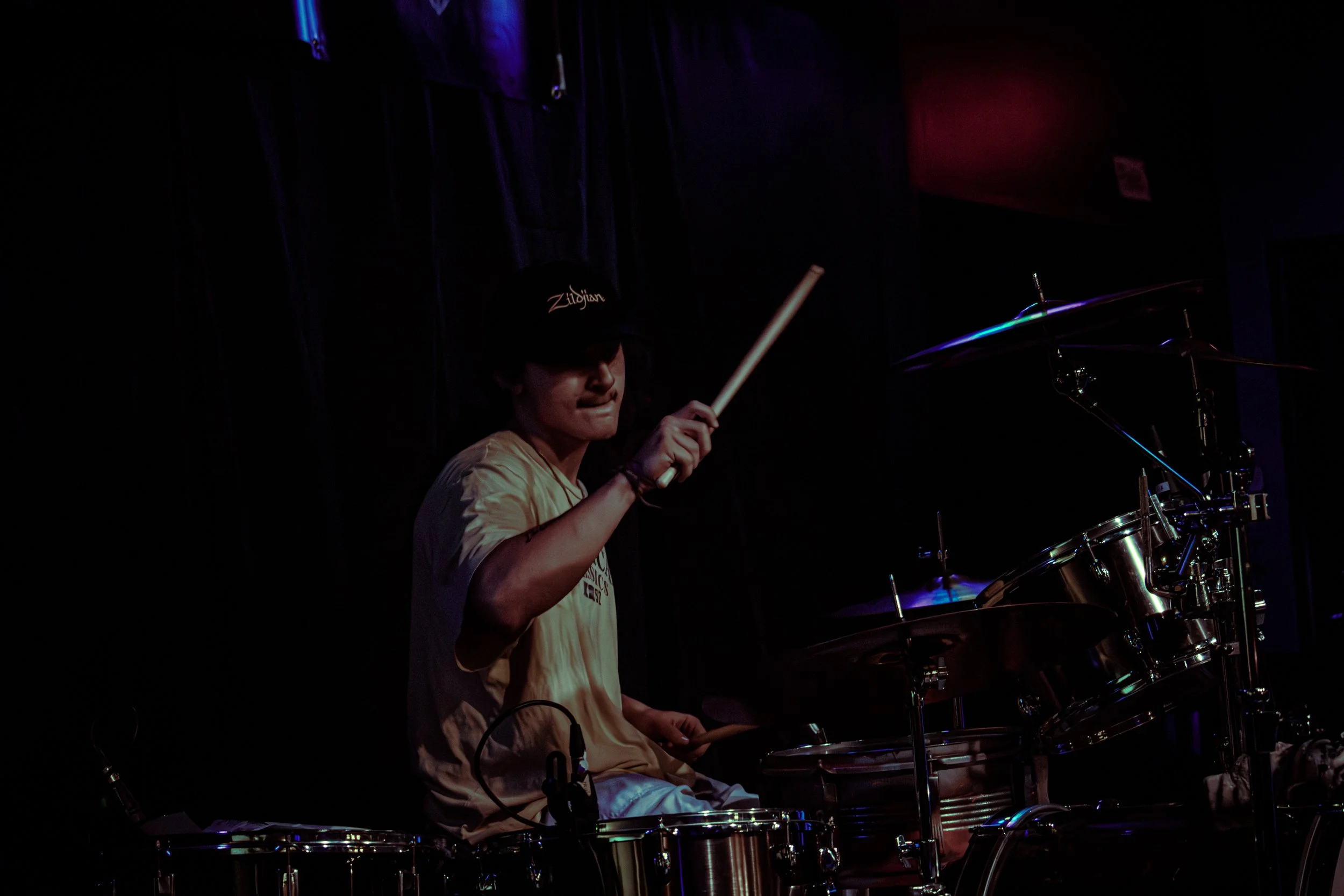 A person playing drums on a dark stage, wearing a black cap and a yellow t-shirt, with a partially visible setting and musical instruments.