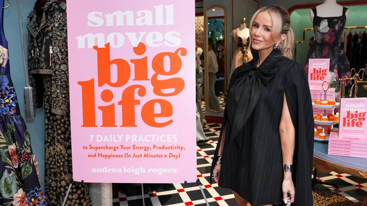 A woman with blonde hair styled in a ponytail wearing a black pinstripe cape dress stands in a boutique with clothing displays. Behind her, a large pink sign reads 'small moves big life' and promotes a book by Andrea Leigh Rogers about daily practices to boost energy, productivity, and happiness.