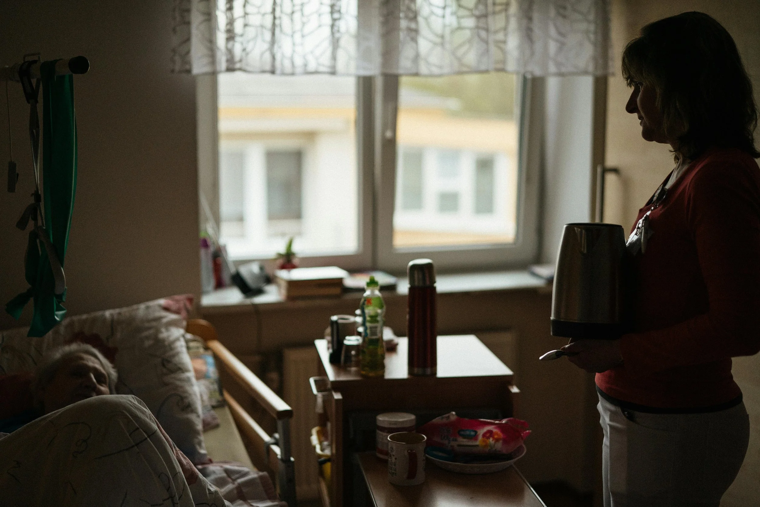 Nurse standing with a coffee pot in a hospital room with a patient lying in bed.