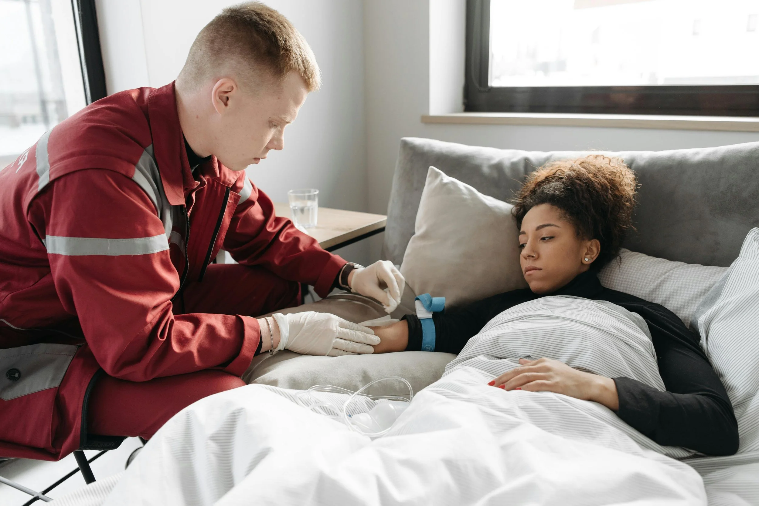 A healthcare worker in a red jacket checks the pulse of a young woman lying in a hospital bed, with a window and a glass of water in the background.
