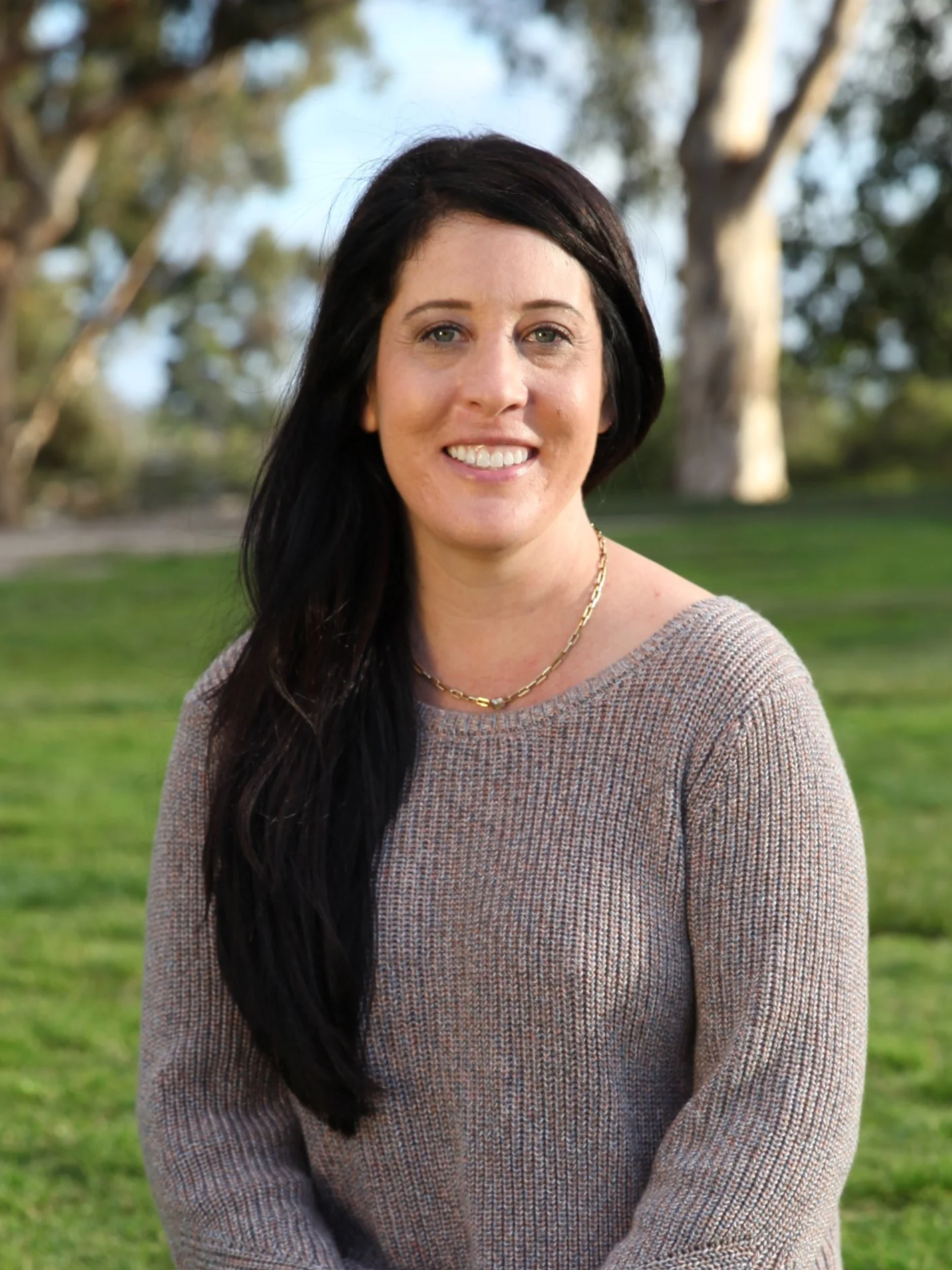 A woman with long dark hair and light skin smiling outdoors in a park with trees and grass.