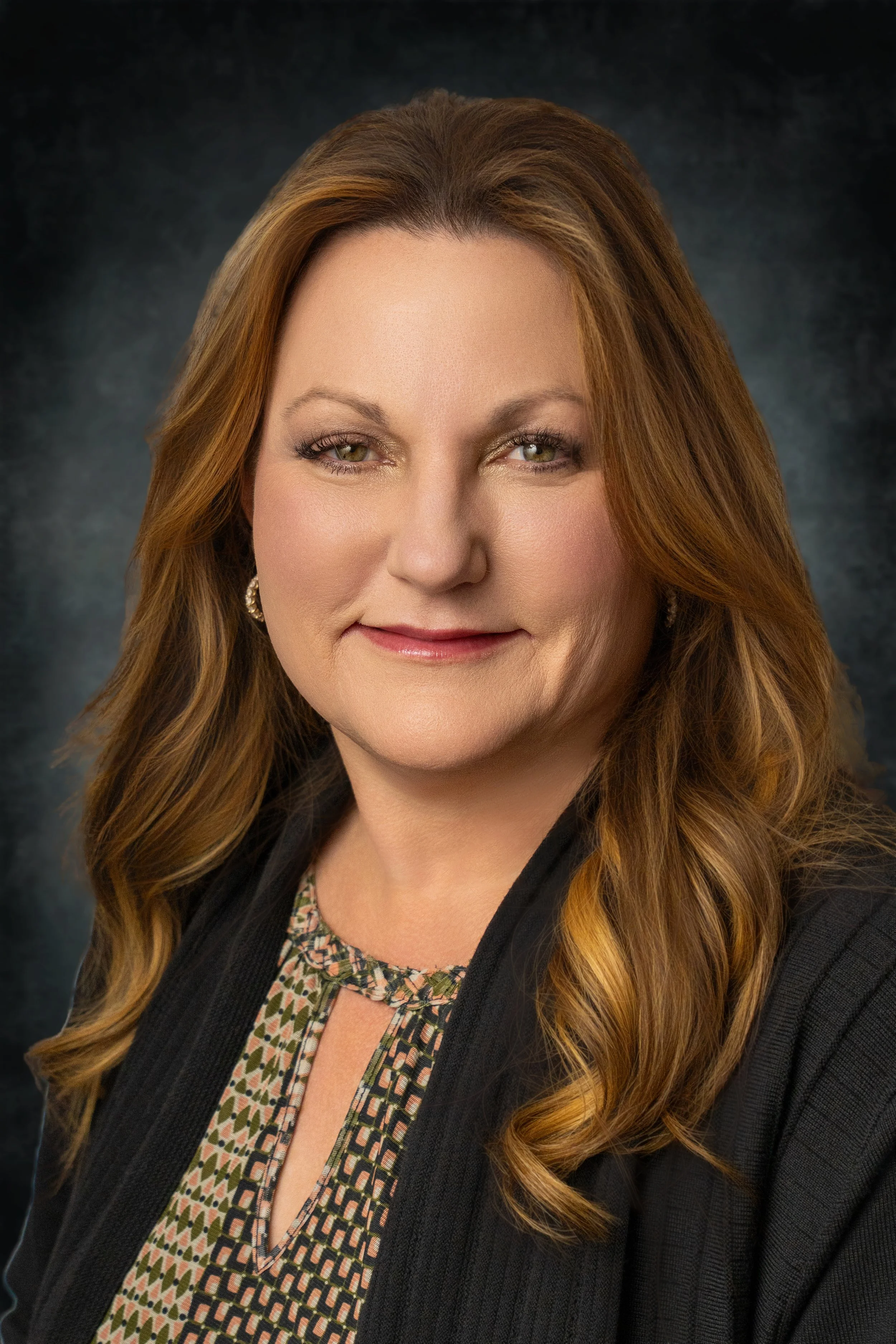 A woman with long, wavy red hair, green eyes, and a light complexion, smiling softly, wearing a black jacket and a patterned blouse, against a dark blurred background.
