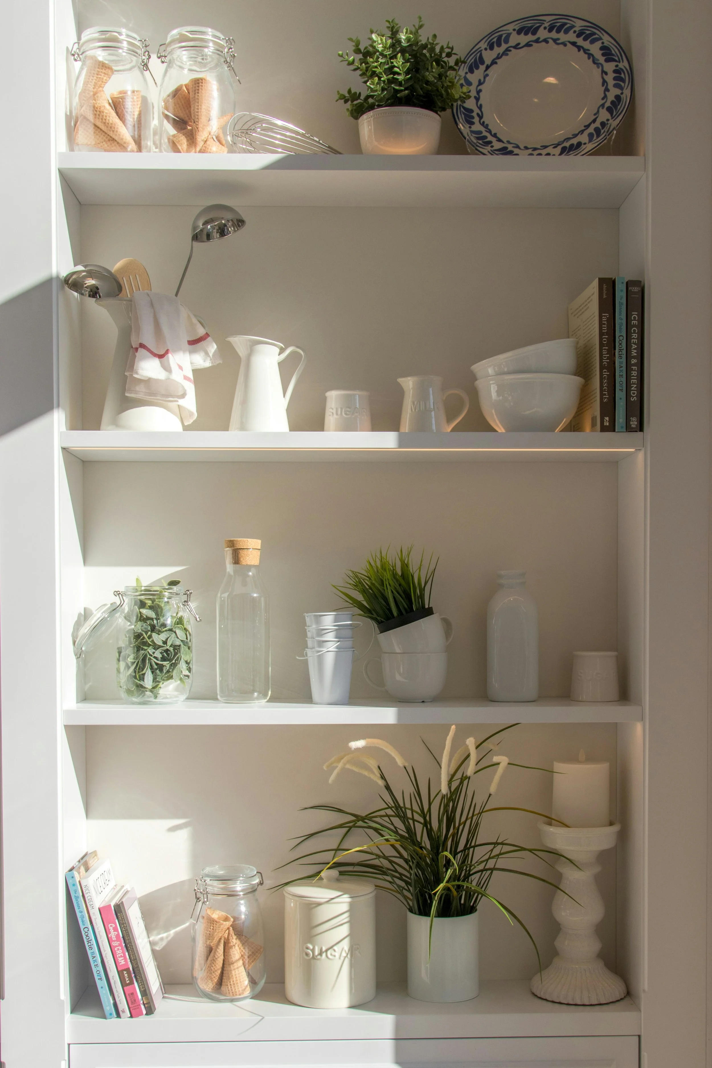 White kitchen shelving unit with glass jars, potted plant, blue-and-white dish, cookbooks, and decorative items including pots, cups, bowls, bottles, and plants.