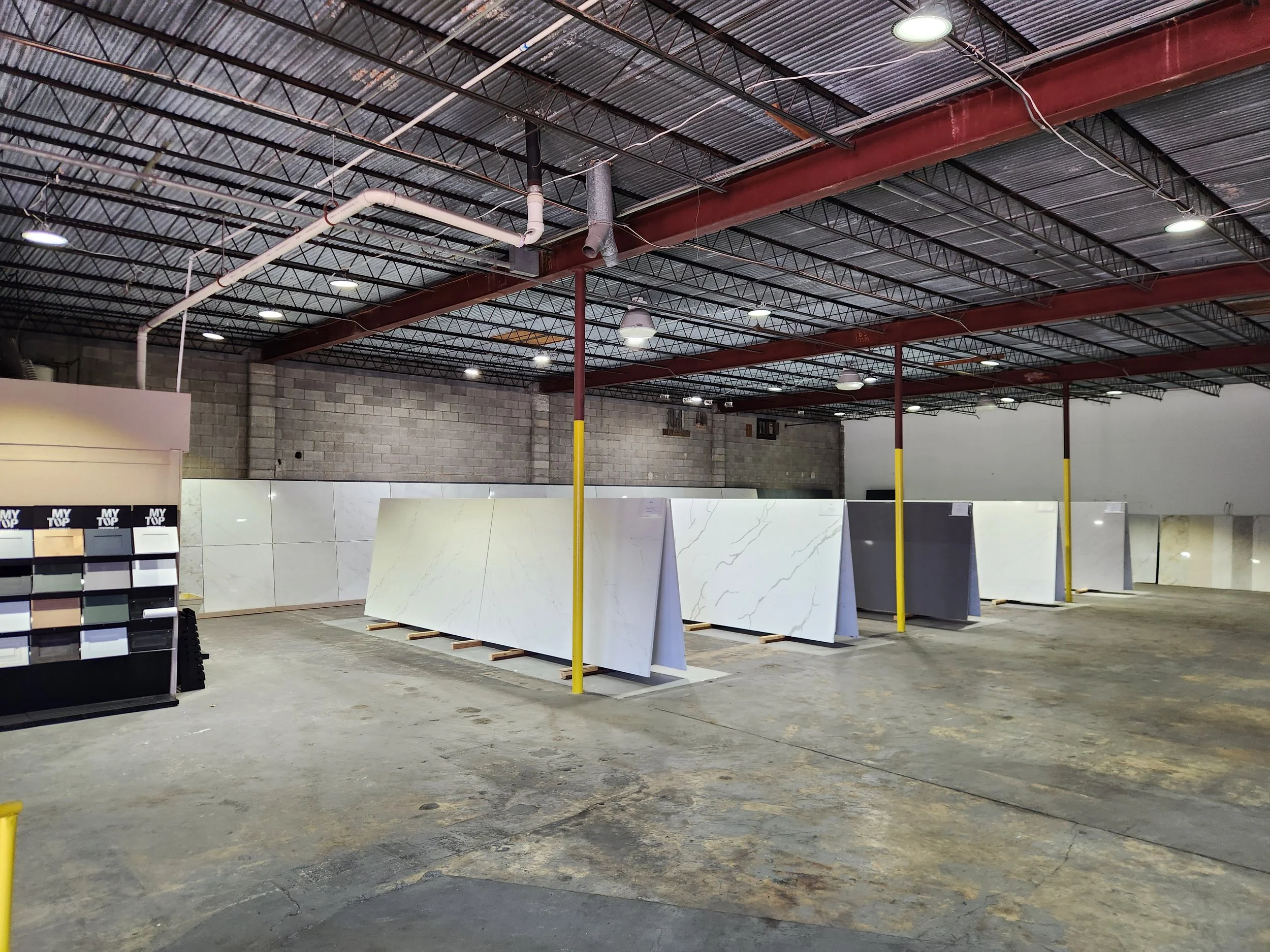 Interior of a warehouse or showroom with large white marble or stone tiles displayed on stands. The space has a high metal ceiling with exposed pipes and lighting fixtures, and chewed concrete floors.