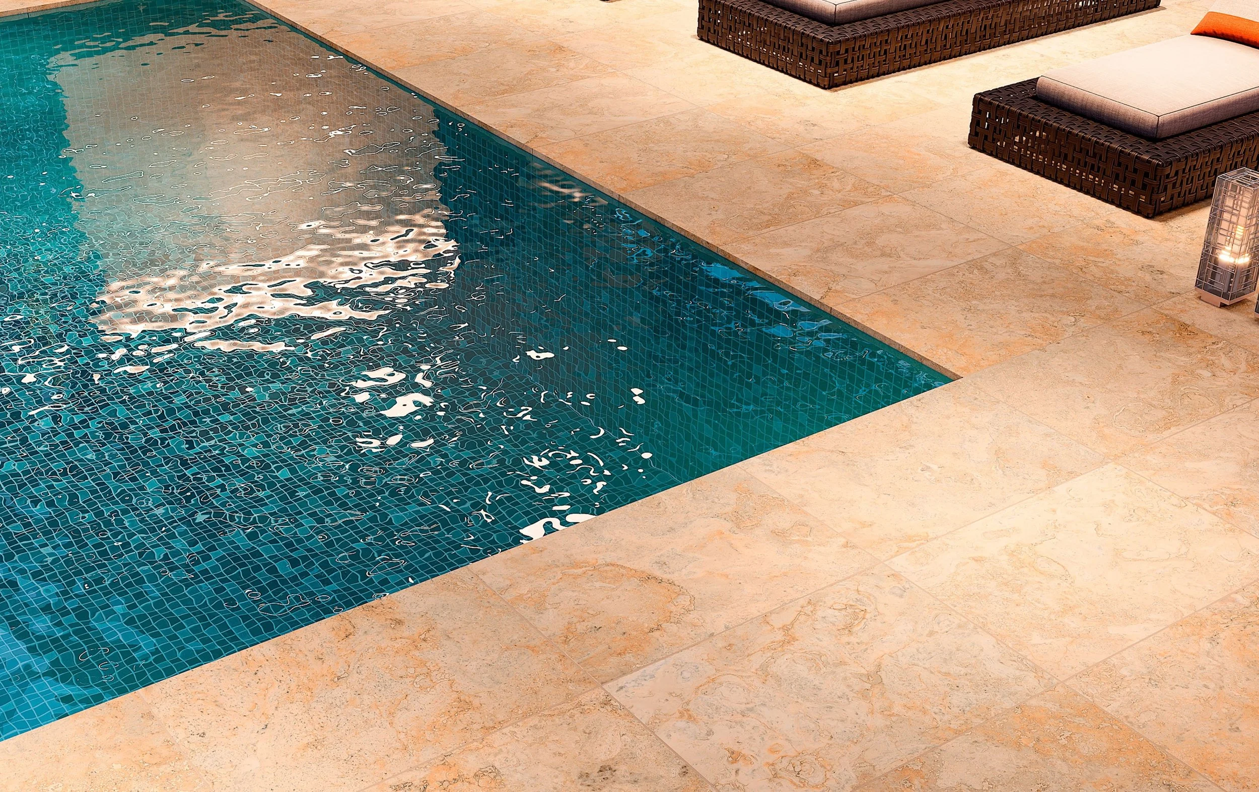 A swimming pool with blue tiles and rippling water, surrounded by beige stone tiles, with wicker lounge chairs and cushions on the deck.