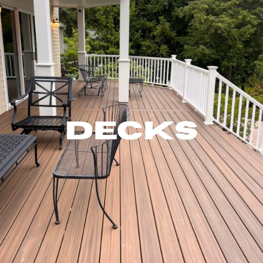 A spacious outdoor deck with wooden flooring, white railing, and outdoor furniture including black metal chairs and a table, surrounded by green trees.