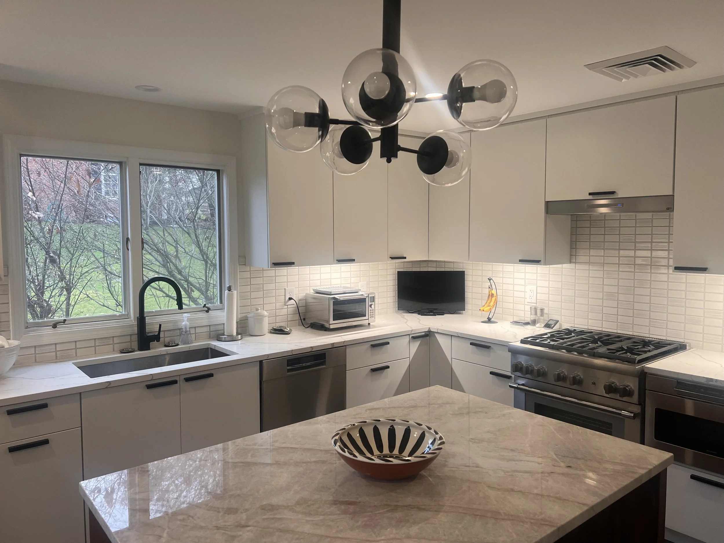 Modern kitchen with white cabinets, marble countertops, a window over the sink, and stainless steel appliances. There is a small TV, a banana-shaped decorative item, a microwave, and a chandelier with spherical glass bulbs hanging from the ceiling.