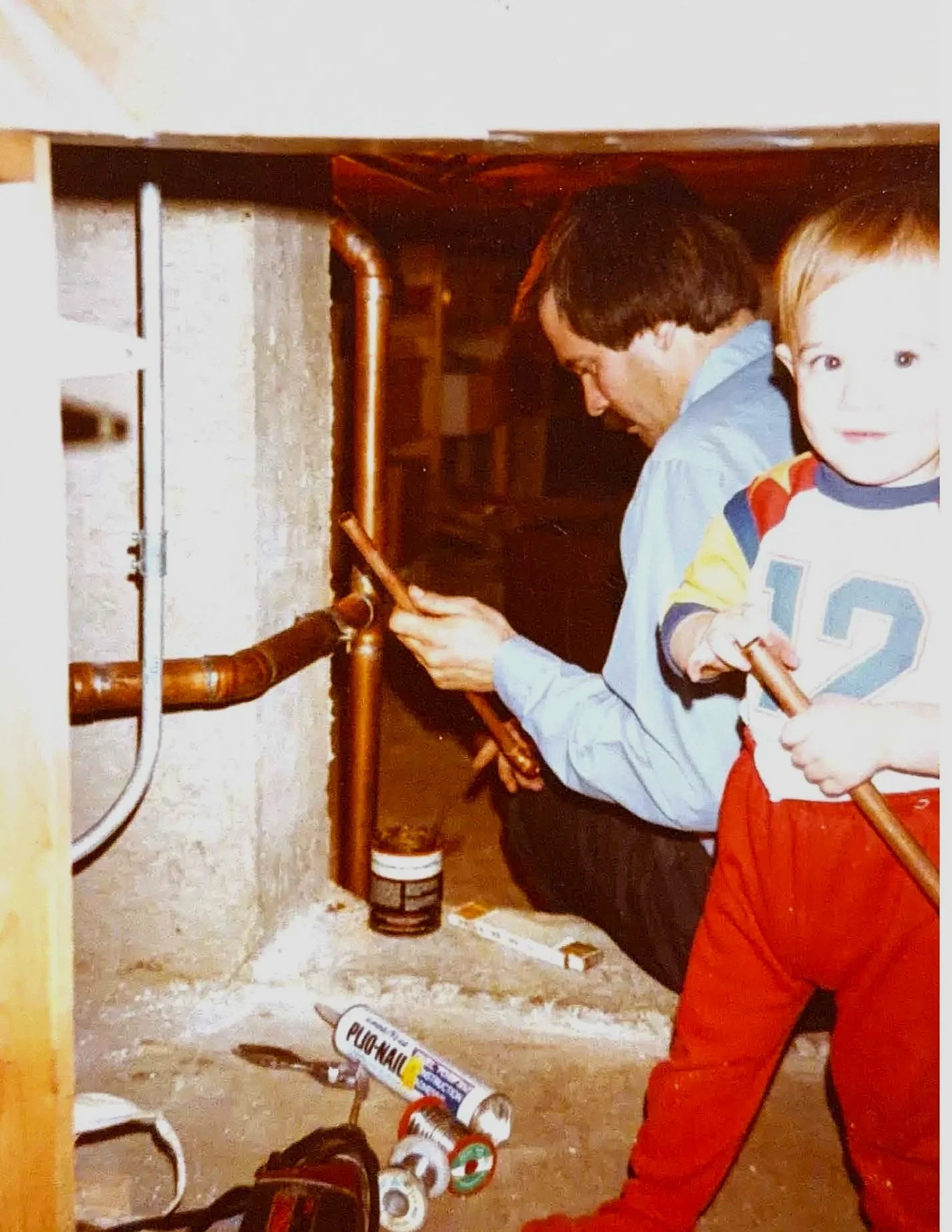 A young boy and a man working on plumbing under a sink, with various tools and supplies on the floor.
