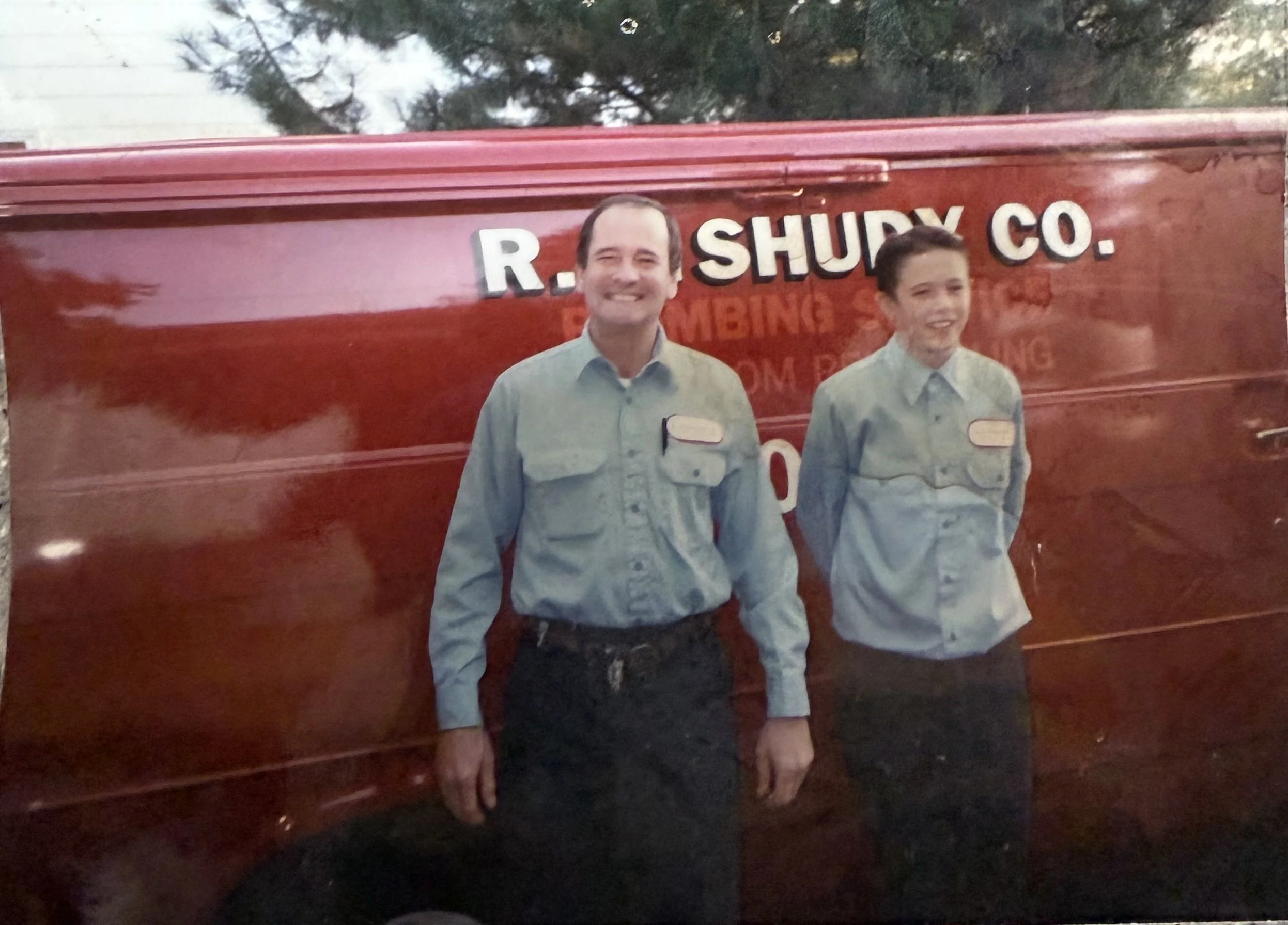 Two people standing in front of a red sign that reads 'R. SHURY CO.' and 'GAMING'. The person on the left is smiling and wearing a light-colored shirt, while the person on the right is also smiling and wearing a similar shirt.