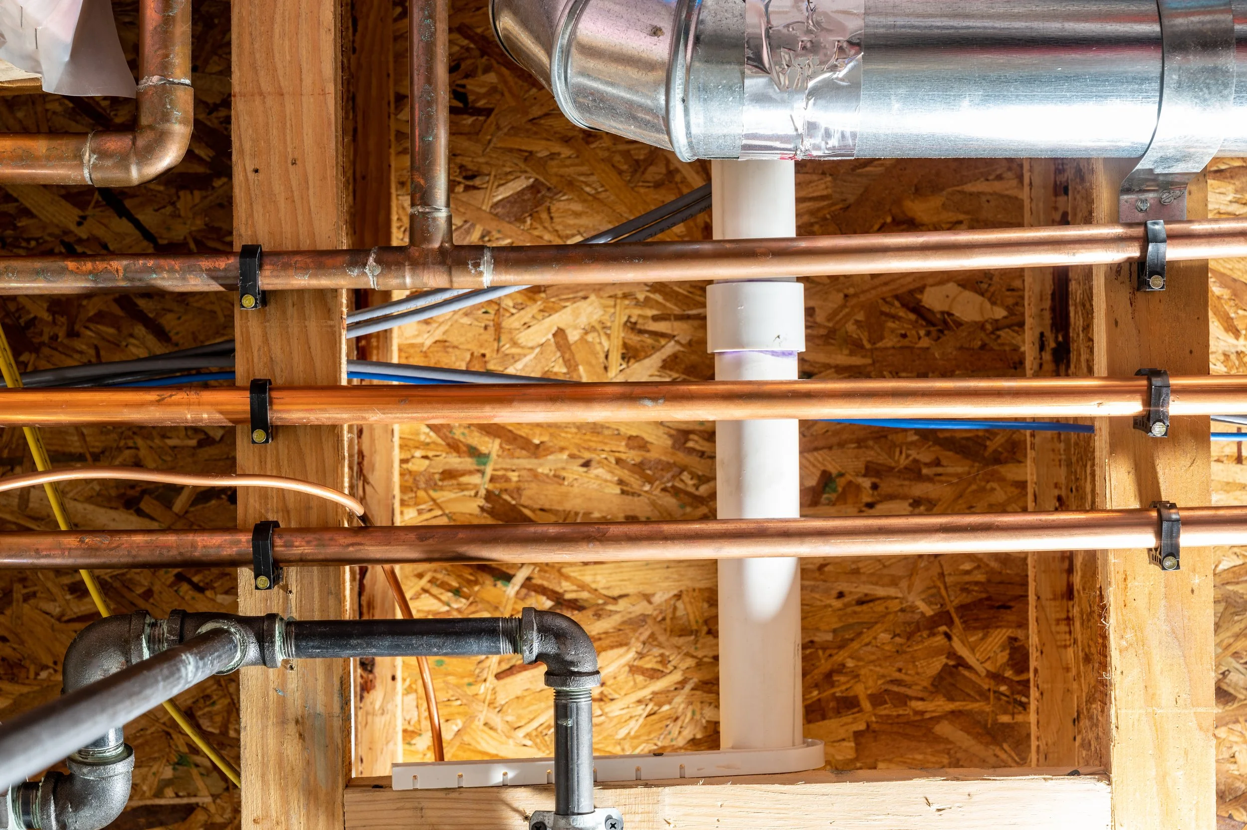 Construction framing with copper and steel pipes, electrical wiring, and a white PVC pipe on a wooden wall.