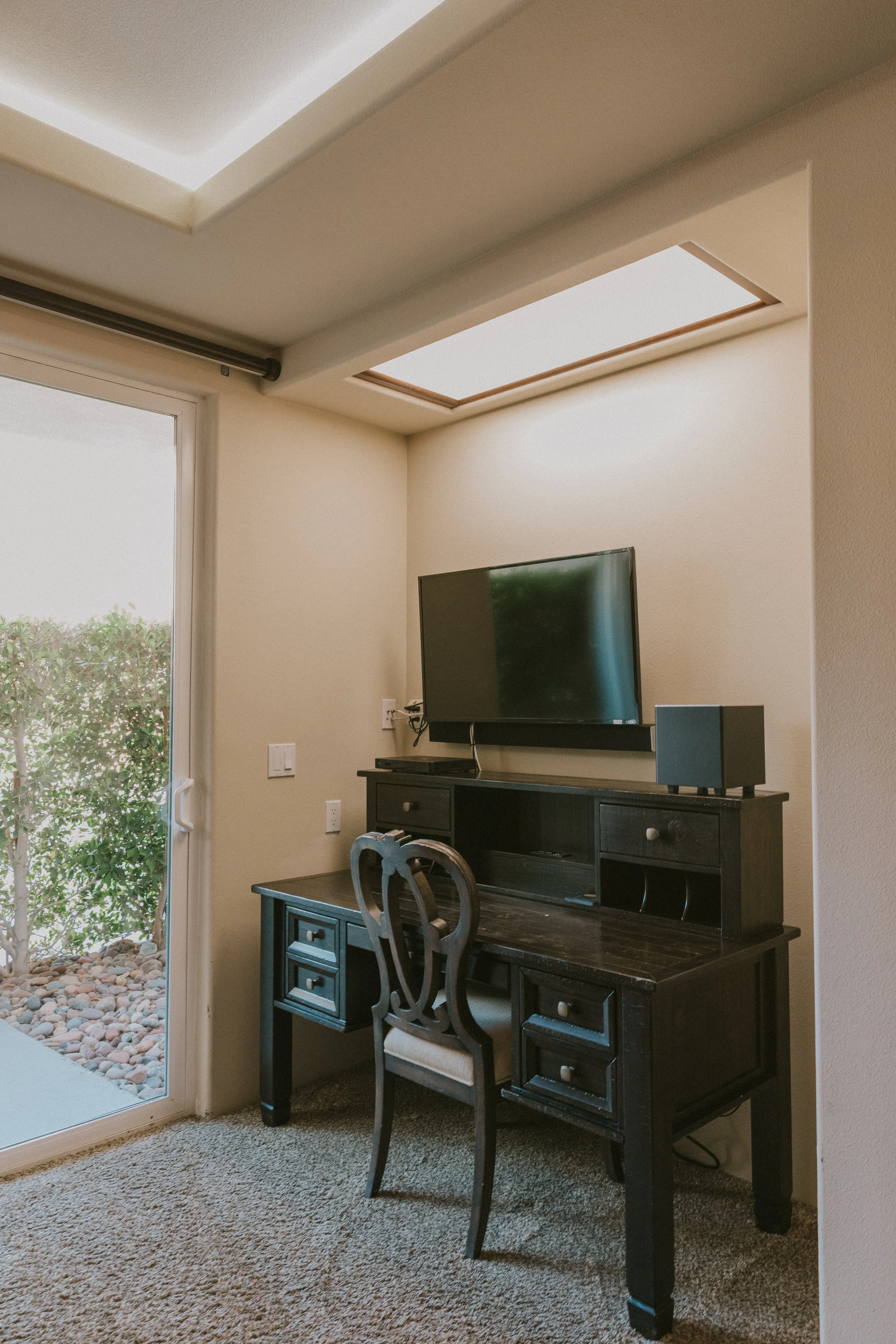 Interior of a room with a black desk, a decorative chair, a wall-mounted TV, and skylights, with a sliding glass door leading outside.