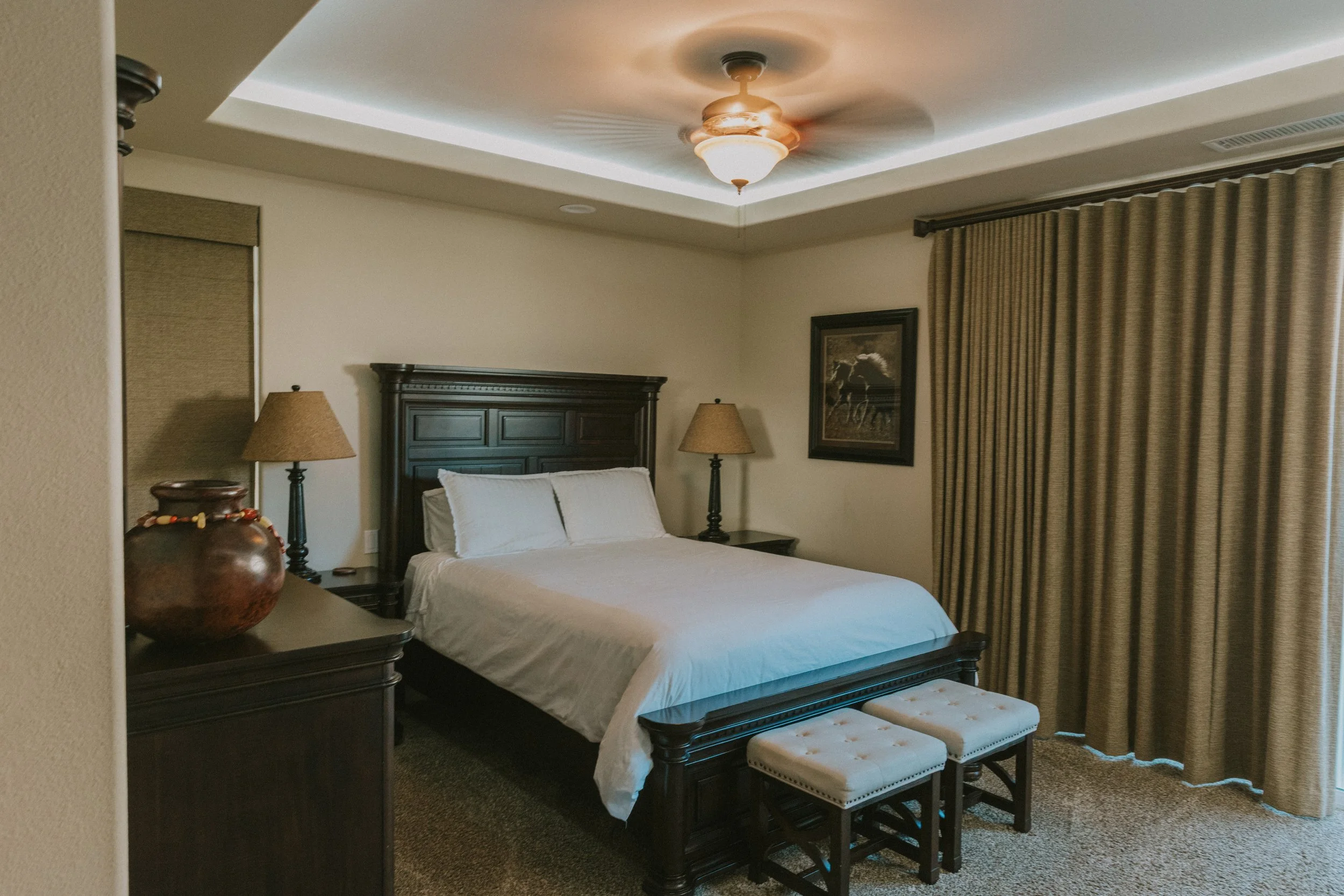A hotel room with a king-sized bed, dark wood headboard, two matching nightstands with lamps, a framed picture on the wall, beige curtains, and a beige ottoman at the foot of the bed. There is a ceiling light fixture and a tray with decorative items 
