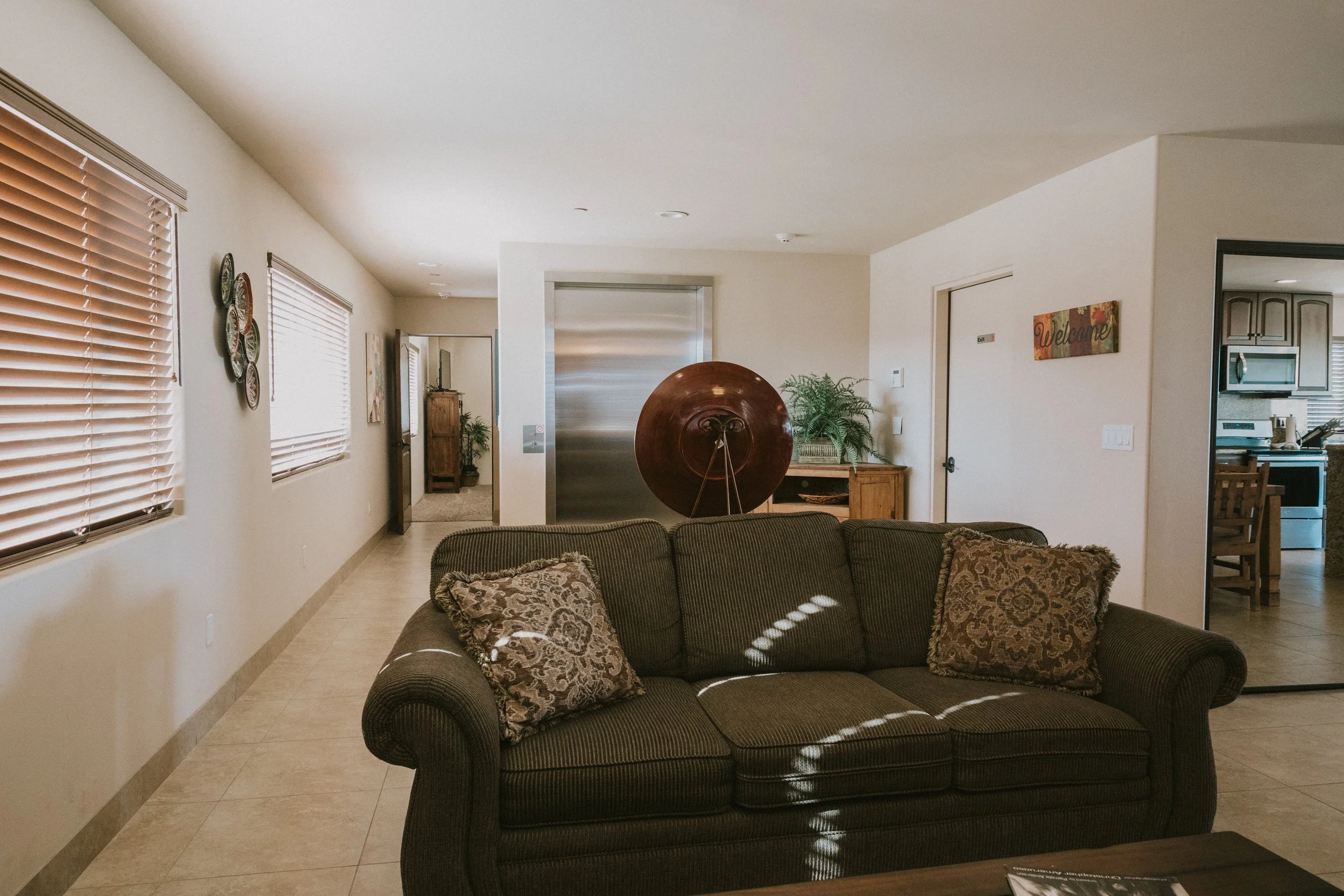Living room with a dark brown couch with decorative pillows, wooden side tables, potted plants, an elevator, and a kitchen area visible through an open doorway.