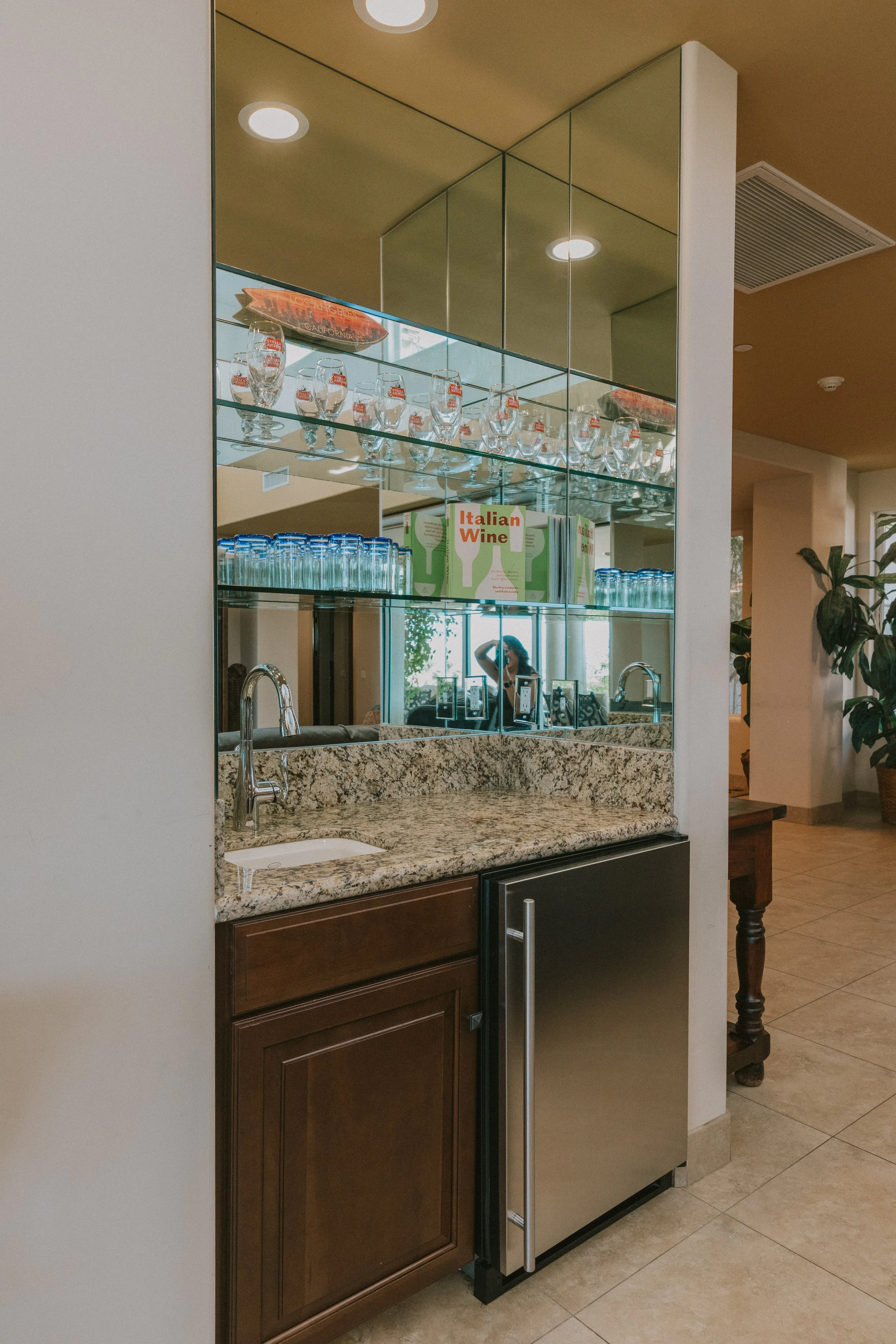 Kitchen area with granite countertop, wooden cabinets, a mini refrigerator, and a mirror reflecting shelves with glasses and a box labeled "Italian Wine."