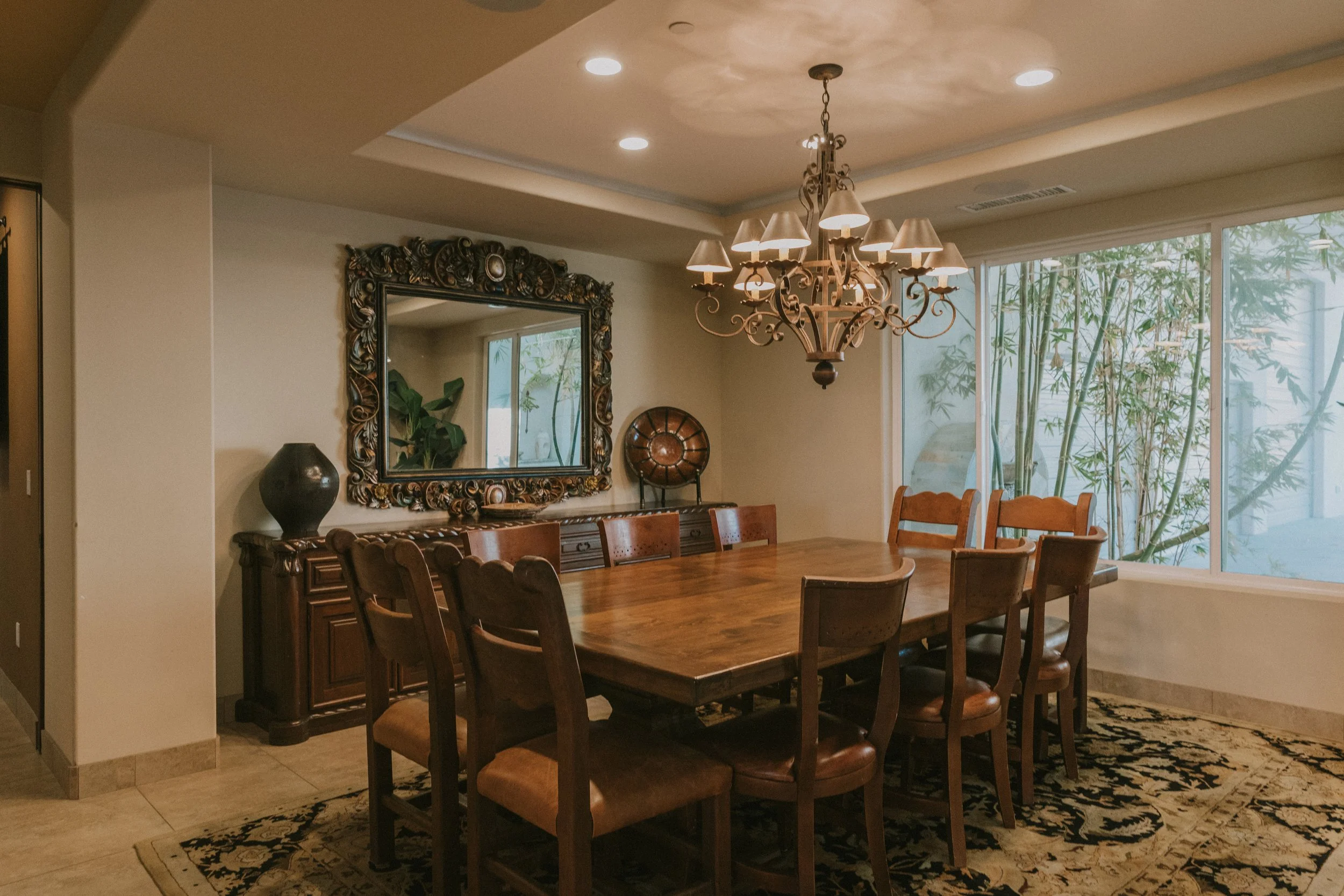 A dining room with a large wooden table surrounded by ten wooden chairs. A ornate chandelier hangs above the table. There is a large mirror and decorative items on a sideboard against the wall. Large window panels show green bamboo outside.