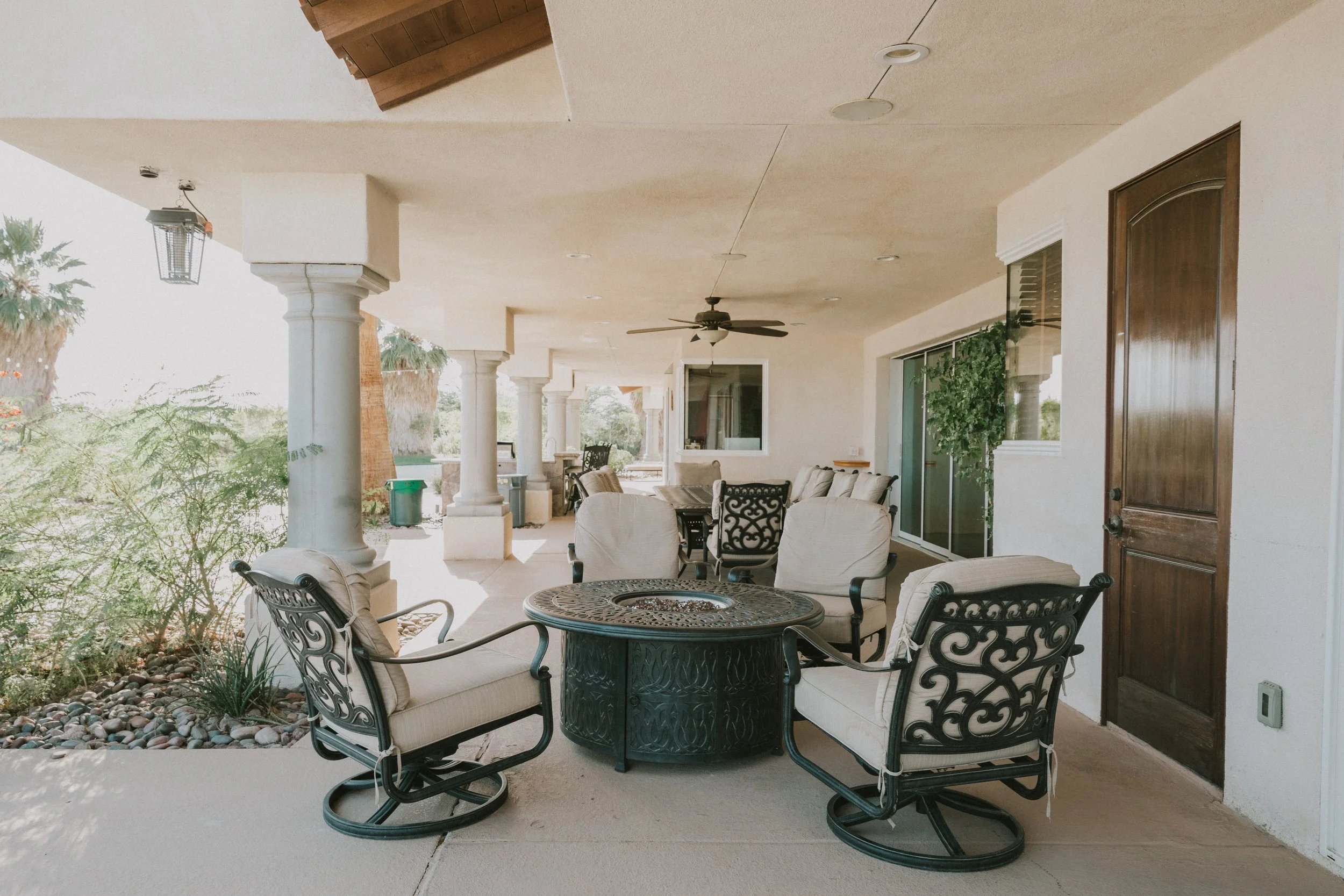 A covered outdoor patio with sitting area, including a round metal table and chairs with beige cushions, ceiling fan, and surrounding columns, with trees and landscape visible beyond.