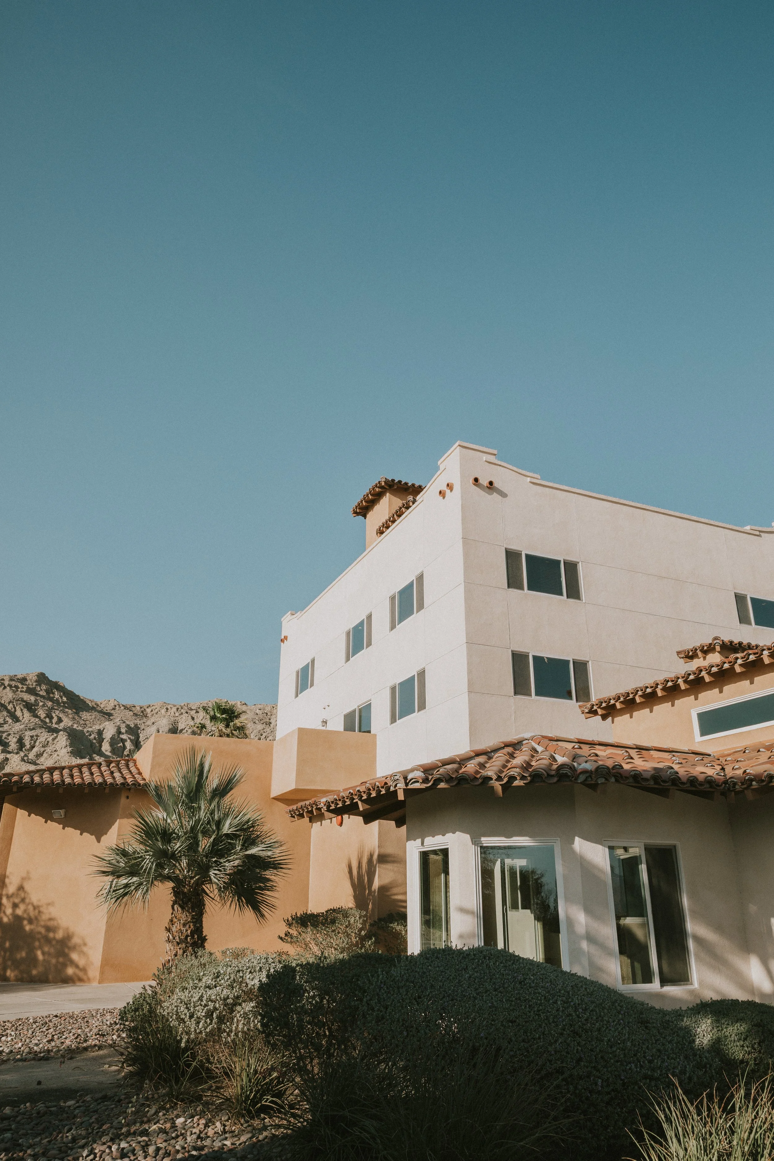 A multi-story modern building with white and beige walls, several windows, and a red-tiled roof, set against a mountainous desert landscape with clear blue sky.