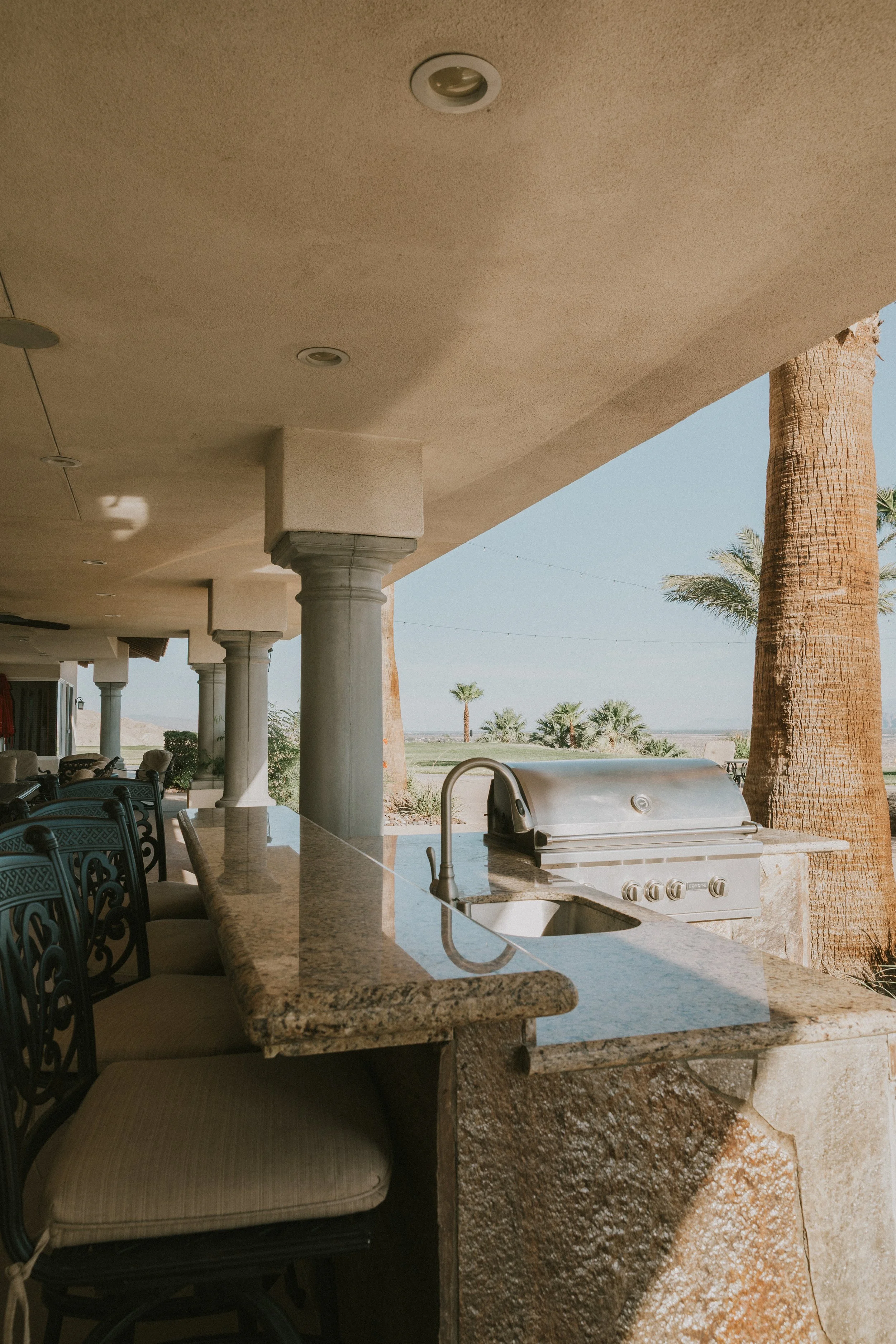 Open outdoor kitchenette with built-in grill, granite countertop, seating, and desert landscape with palm trees in background.