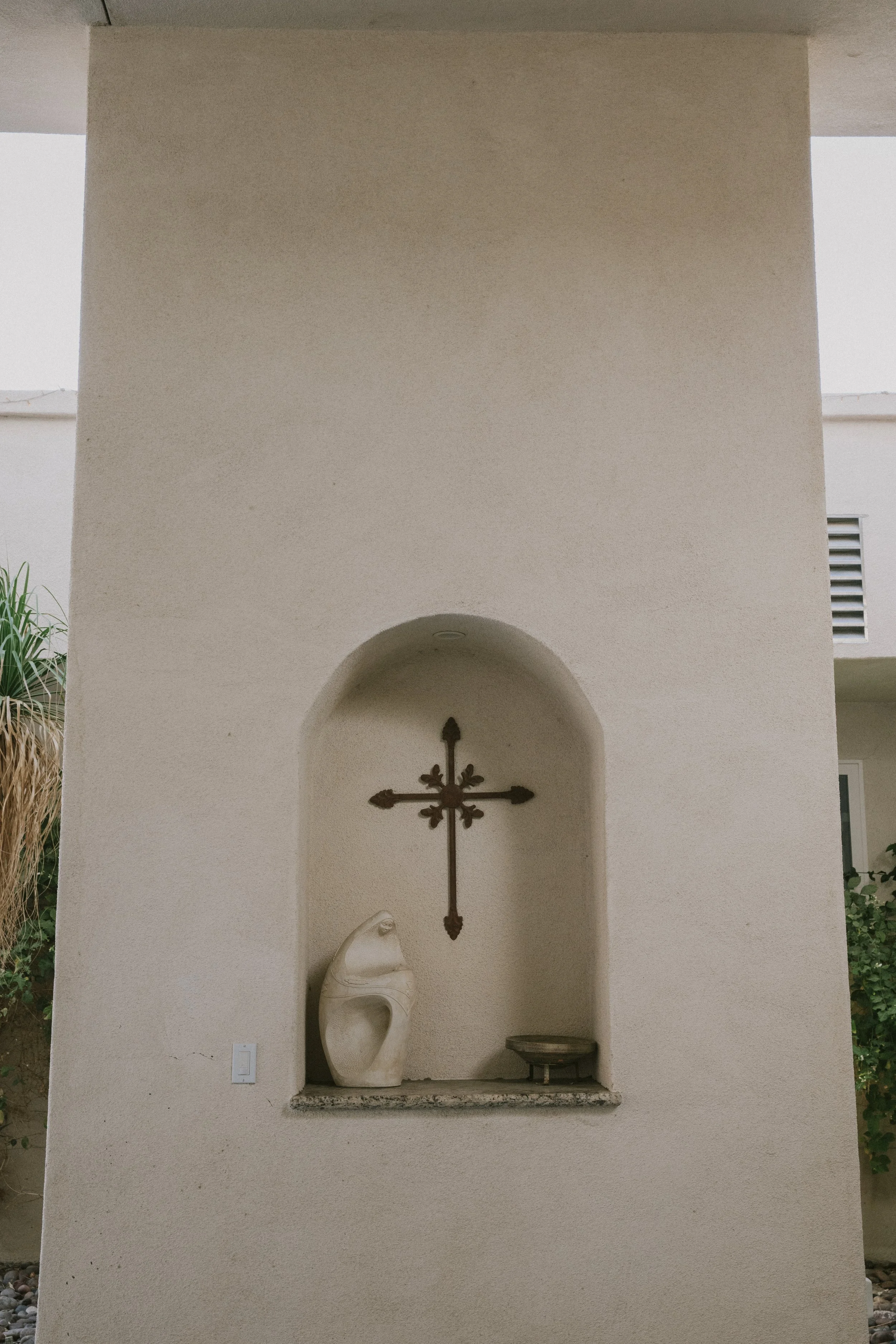 A white carved sculpture of a mermaid in a small alcove with a metal cross hanging on the wall above it. There's a small platform beneath the sculpture and a small bowl or dish to the right.
