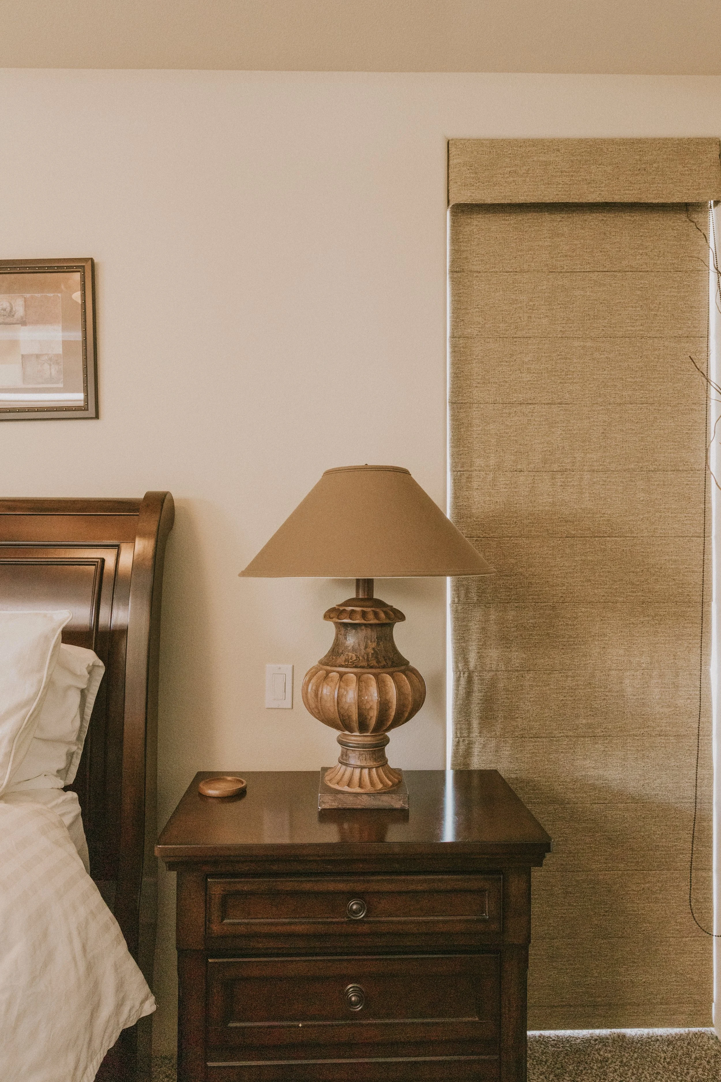 A wooden bedside table with three drawers, ornamented with a decorative lamp with a beige shade, situated next to a bed with white bedsheets, beside a window with beige textured curtains and a framed photo on the wall.