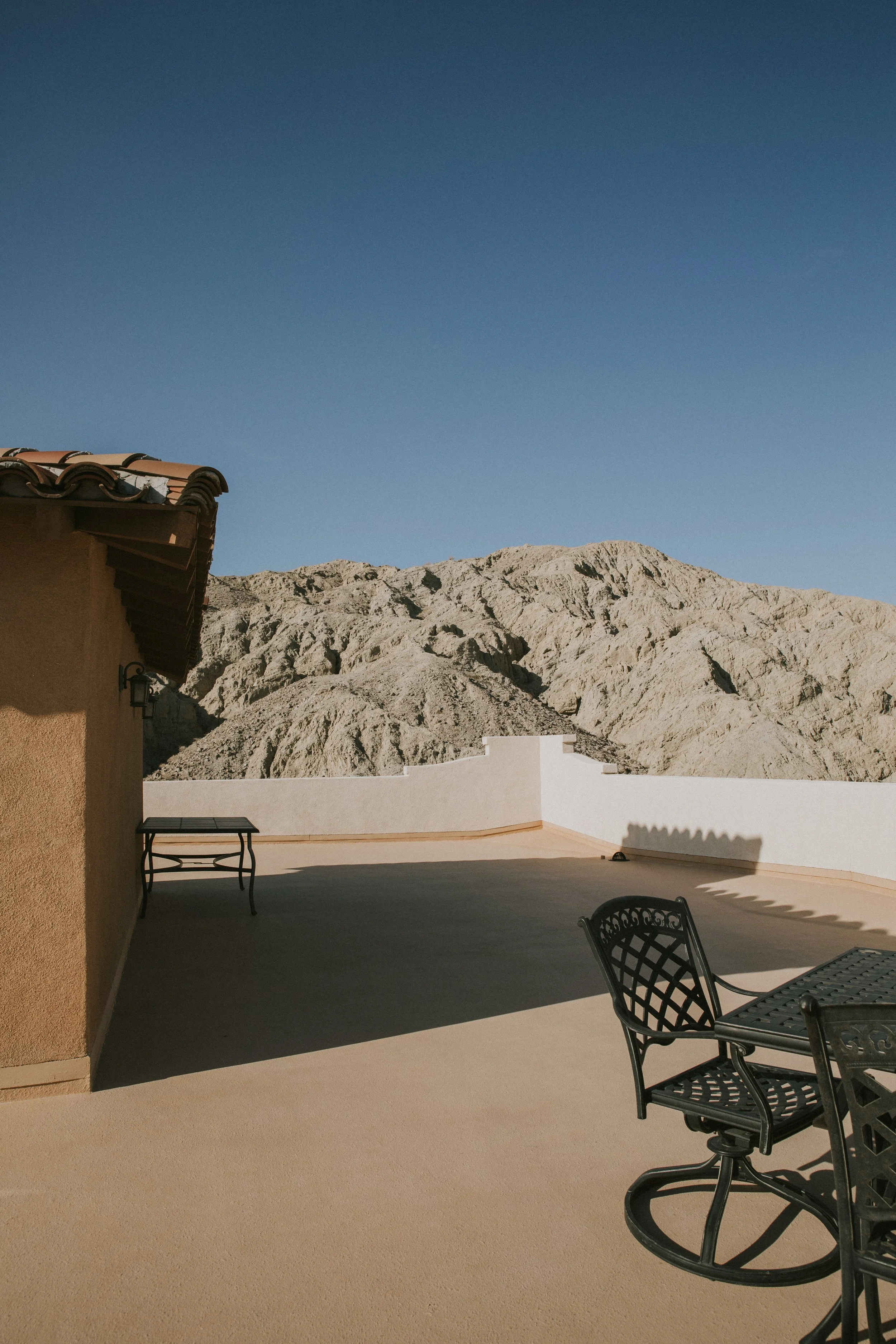 An outdoor patio with black metal chairs alongside a beige stucco wall, overlooking rugged gray mountain terrain under a clear blue sky.