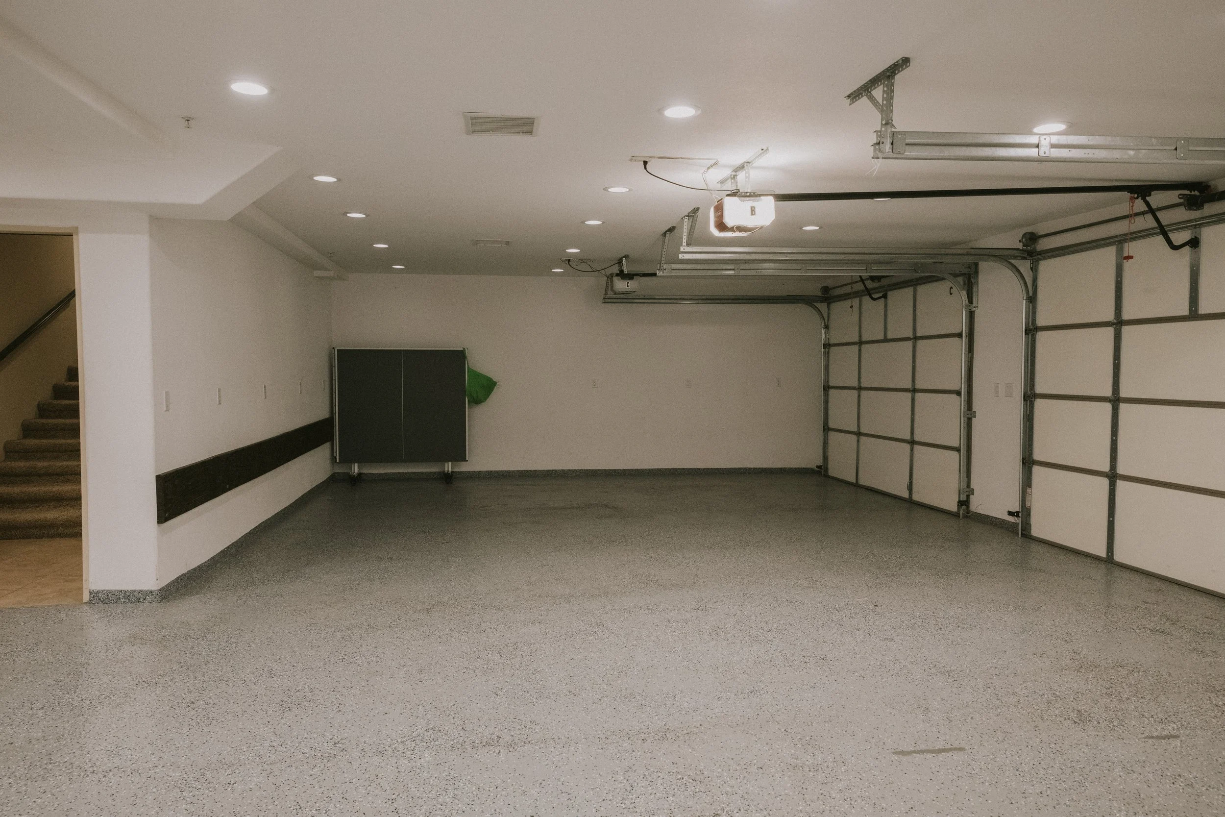 Empty garage with white walls, gray speckled floor, overhead lighting, and a partially open garage door.