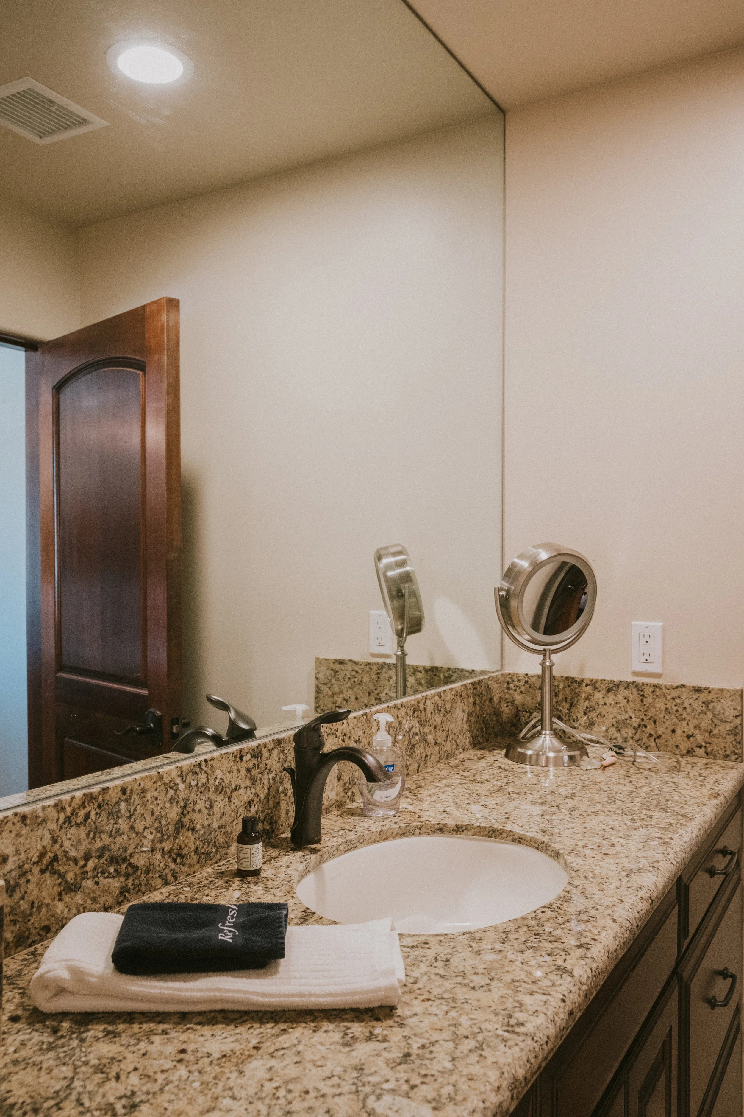 A bathroom countertop with a mirror, a makeup mirror, a soap dispenser, a small bottle, and neatly folded towels with 'Refreshed' embroidered on one in black. The countertop is granite, with a sink and a black faucet. The background shows a wall with