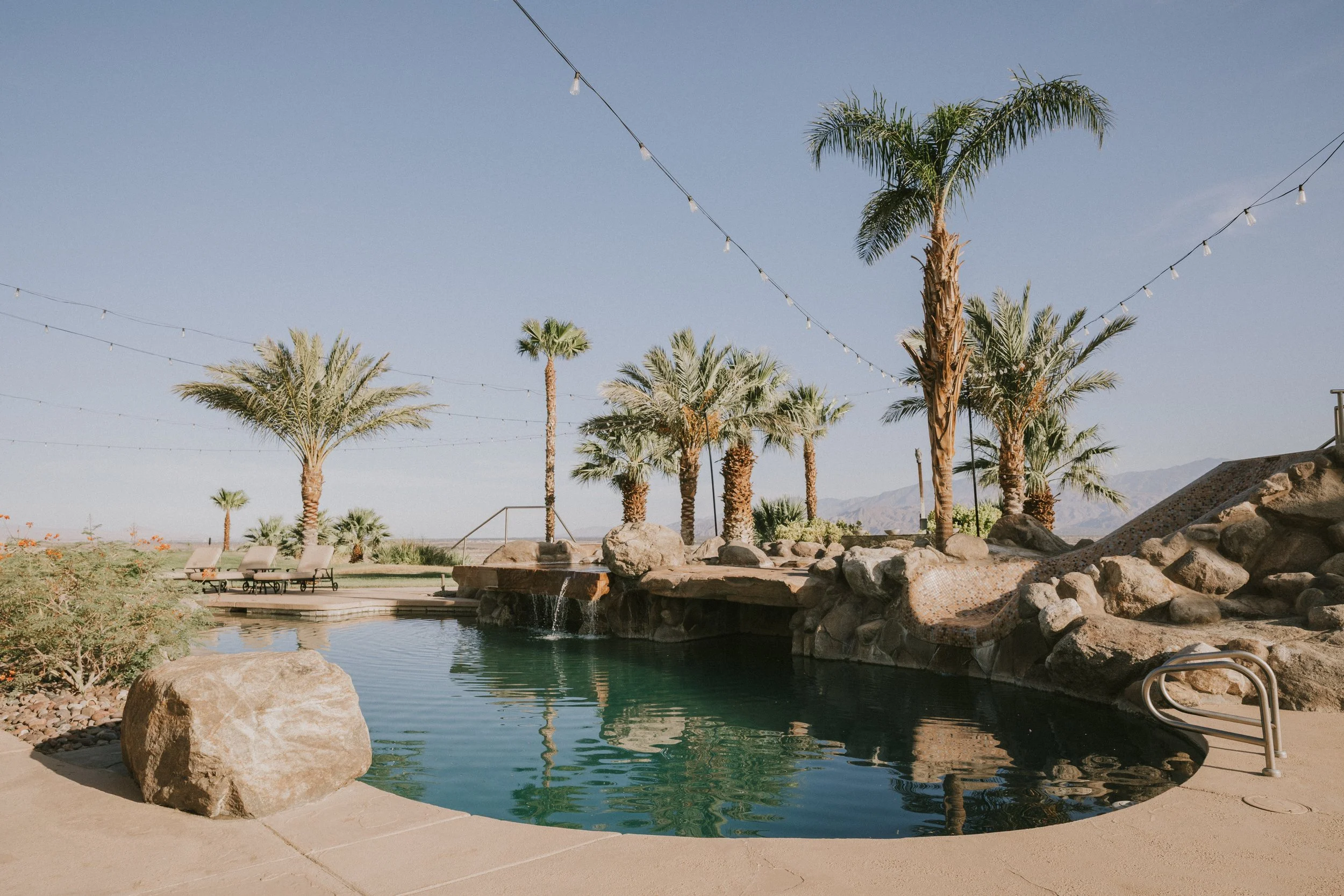 Swimming pool with rocks and palm trees decorated with string lights in a desert or mountain area.