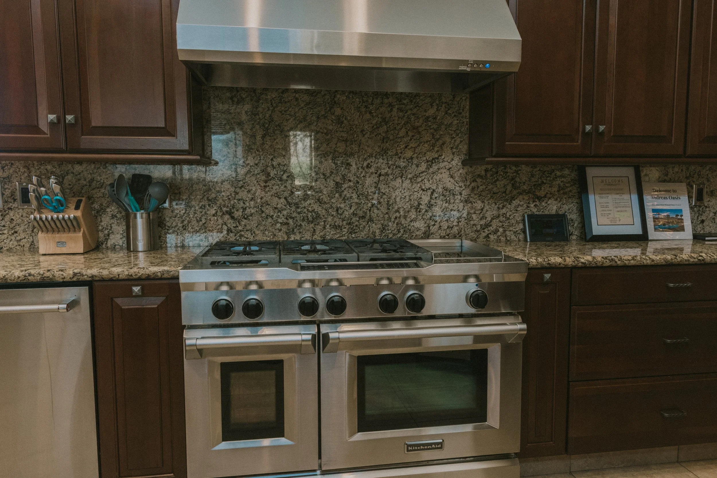 Kitchen with granite countertop, brown cabinets, stainless steel stove and range hood, and framed welcome signs.