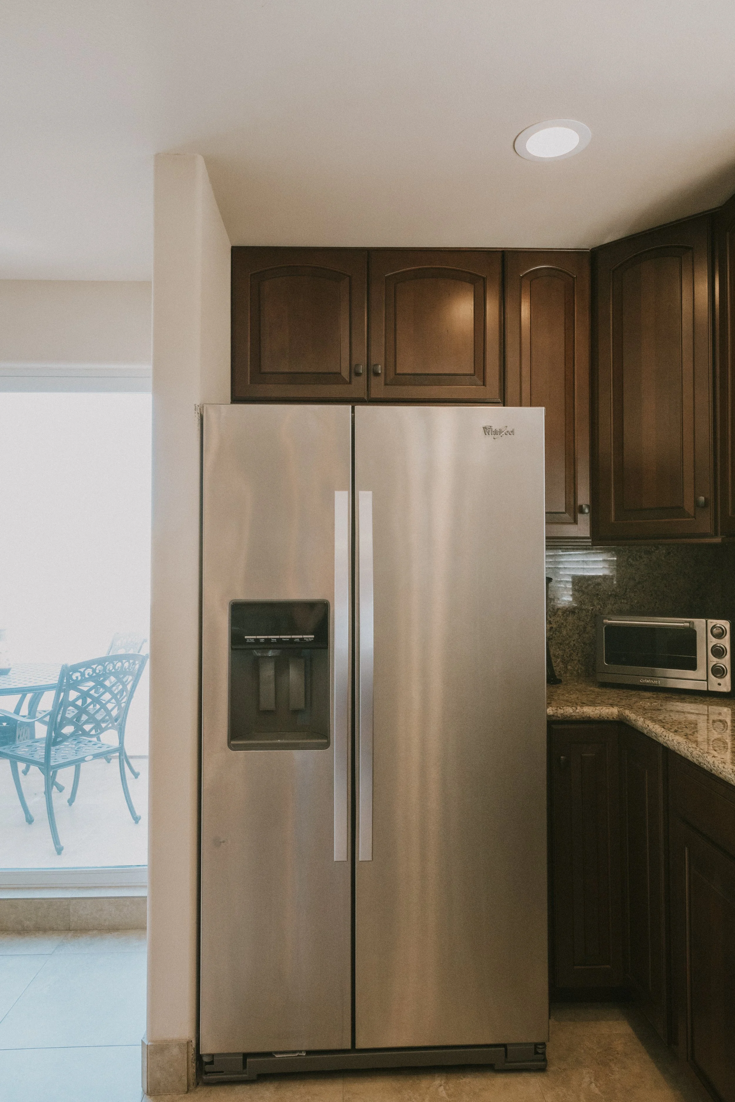 A stainless steel, side-by-side refrigerator with an ice and water dispenser in a kitchen with dark wooden cabinets and beige granite countertop.