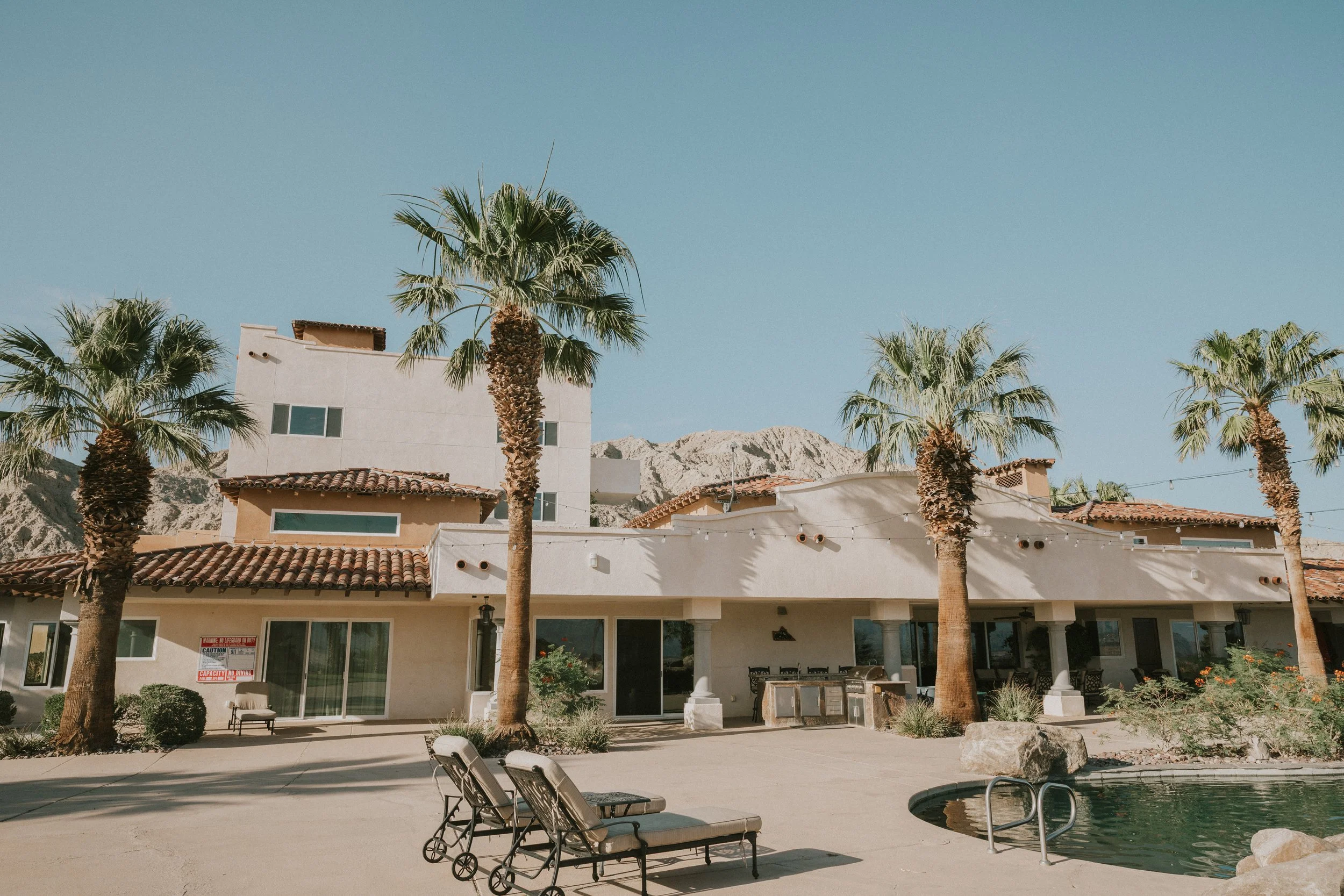 A residential building with palm trees in front, a swimming pool with lounge chairs, and mountains in the background under a clear blue sky.