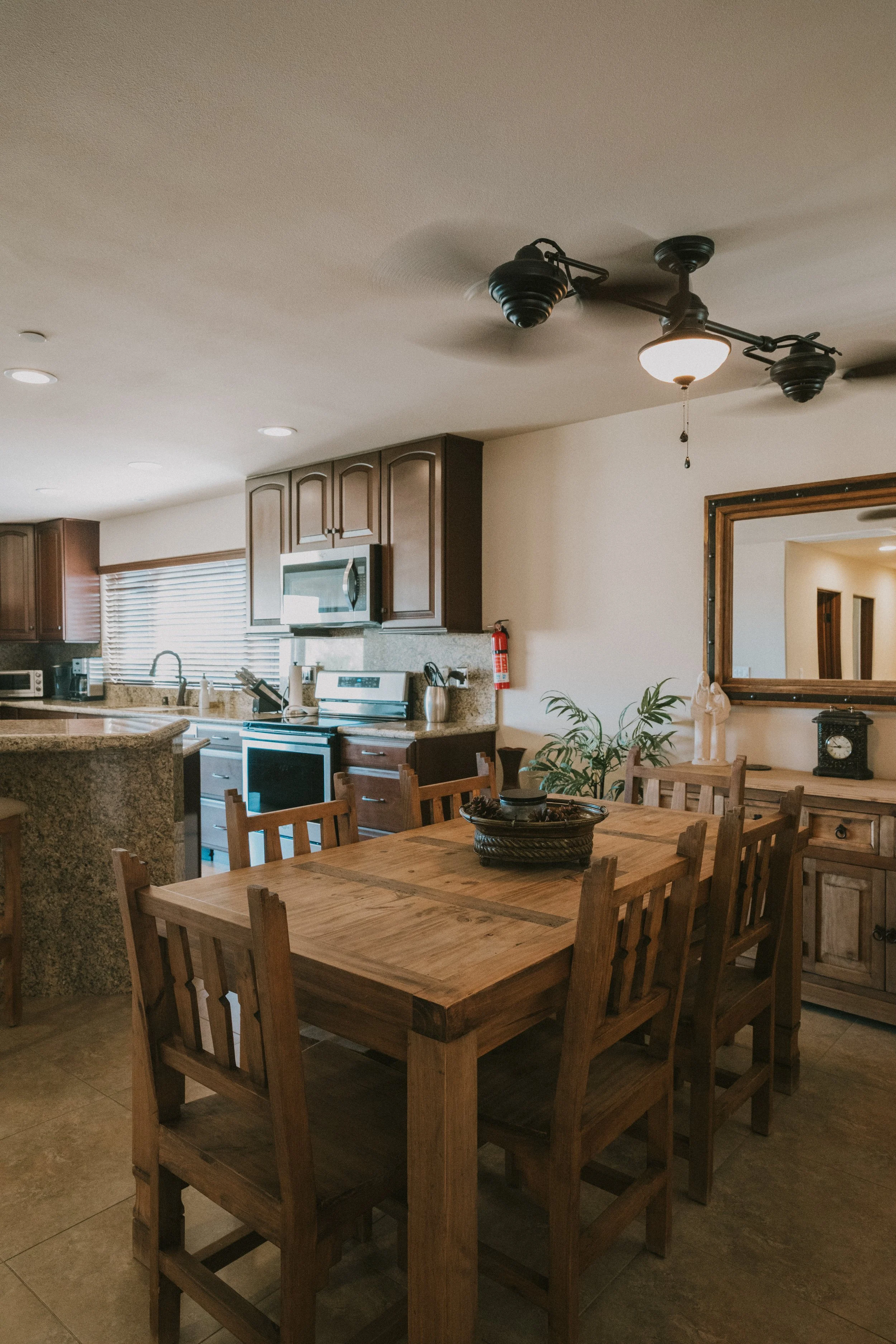 Kitchen with wooden dining table and chairs, kitchen appliances, cabinets, window with blinds, and a framed mirror on the wall.