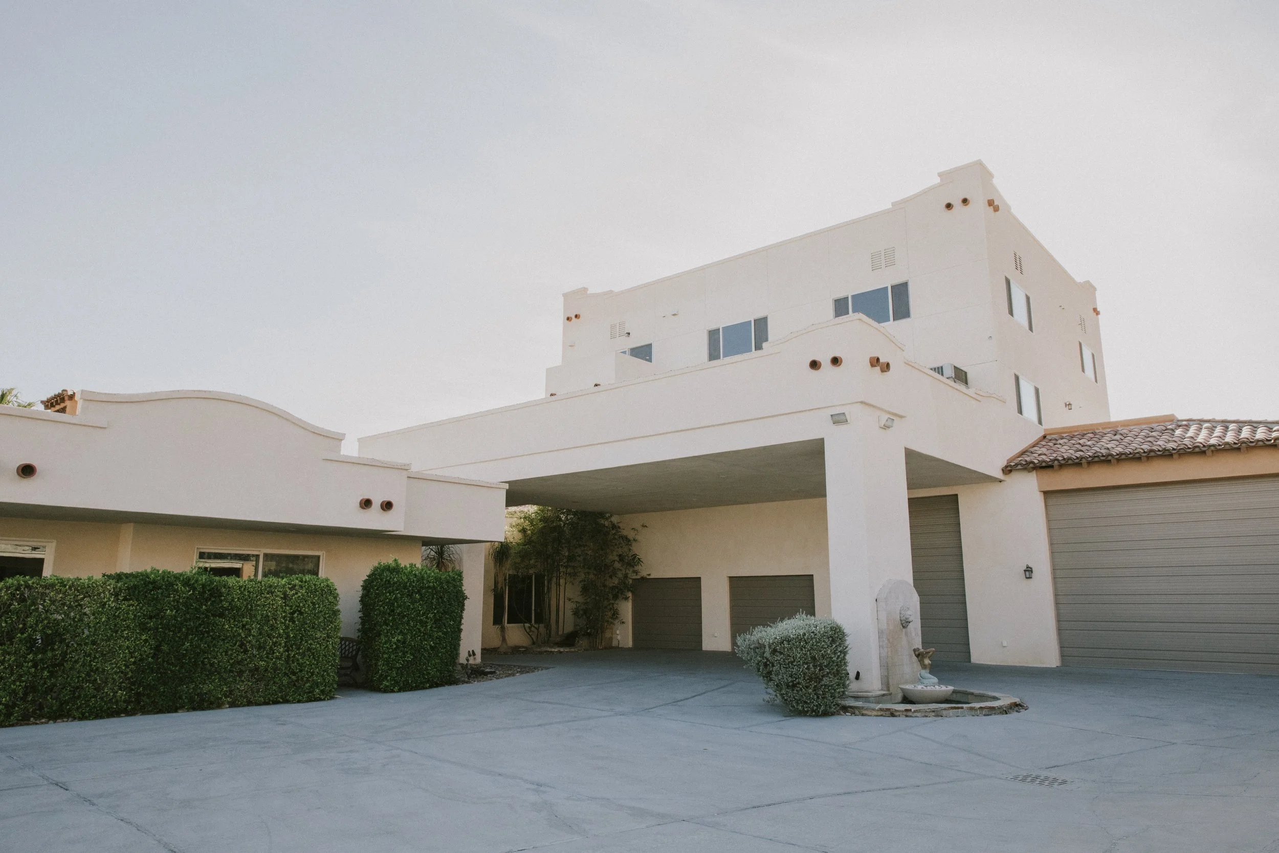 A modern white multi-story house with a tiled roof and multiple windows, surrounded by a driveway, green bushes, and small trees.