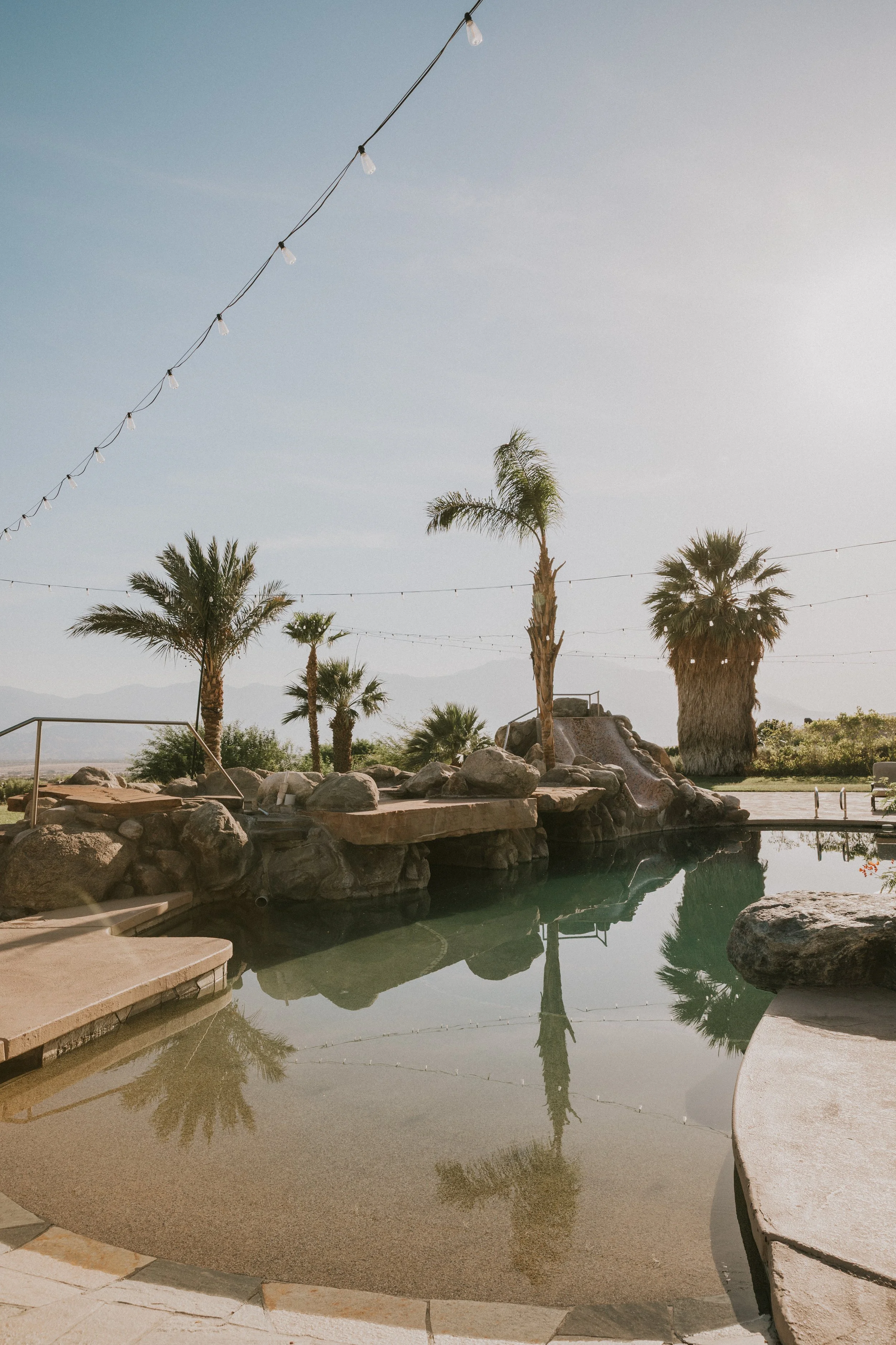 Palm trees around a small outdoor pool with rocks and stone features, string lights overhead, with mountains in the background on a clear day.