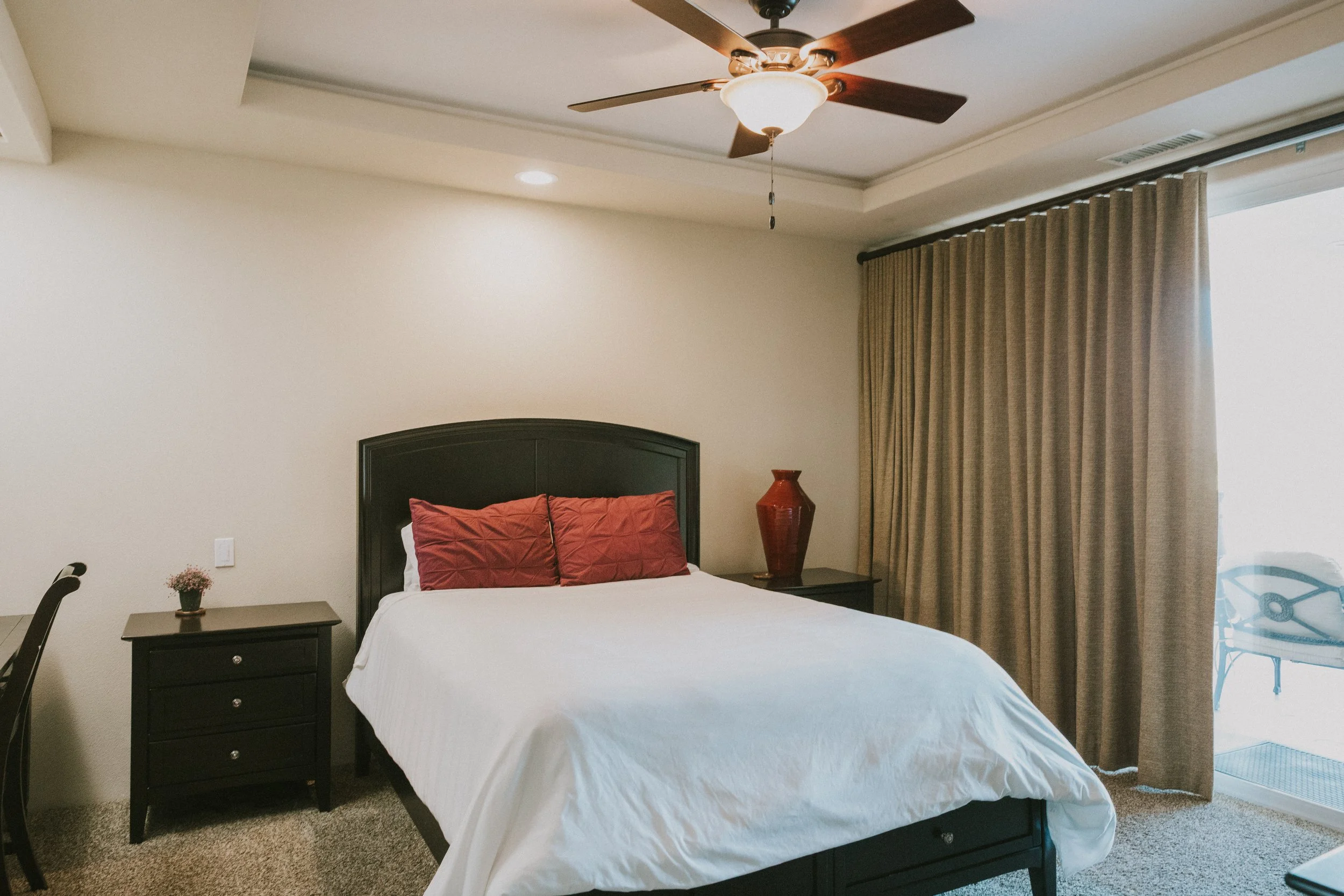 A bedroom with a large bed with white bedding and two red pillows, a black headboard, and a matching black nightstand with a small potted plant. A red vase is on the nightstand. There's a ceiling fan with wooden blades and a light, beige curtains, an