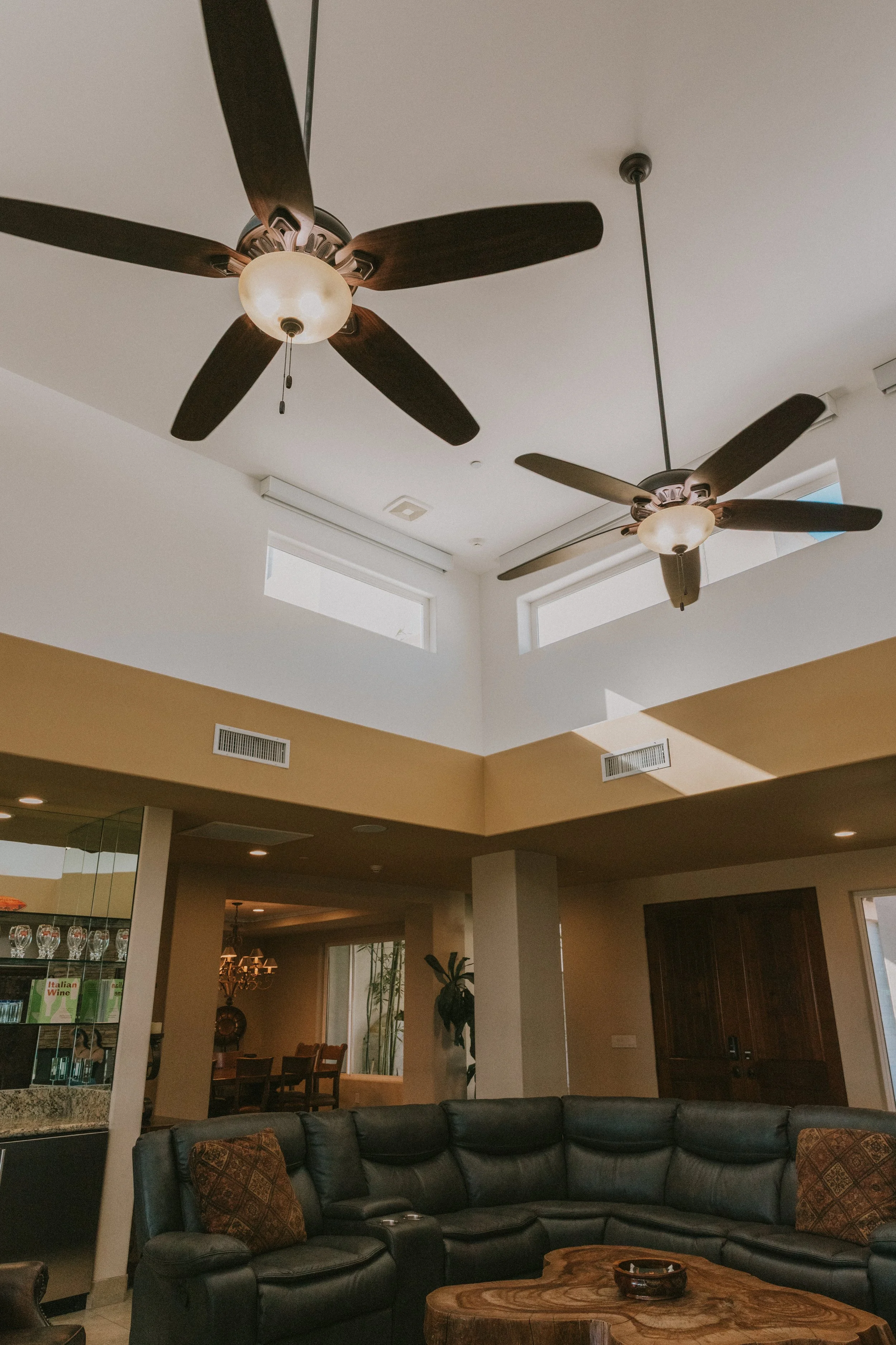 Living room with two ceiling fans with dark wood blades and frosted glass light covers, high ceilings, and large windows, with a dark leather sectional sofa and a wooden coffee table in front.