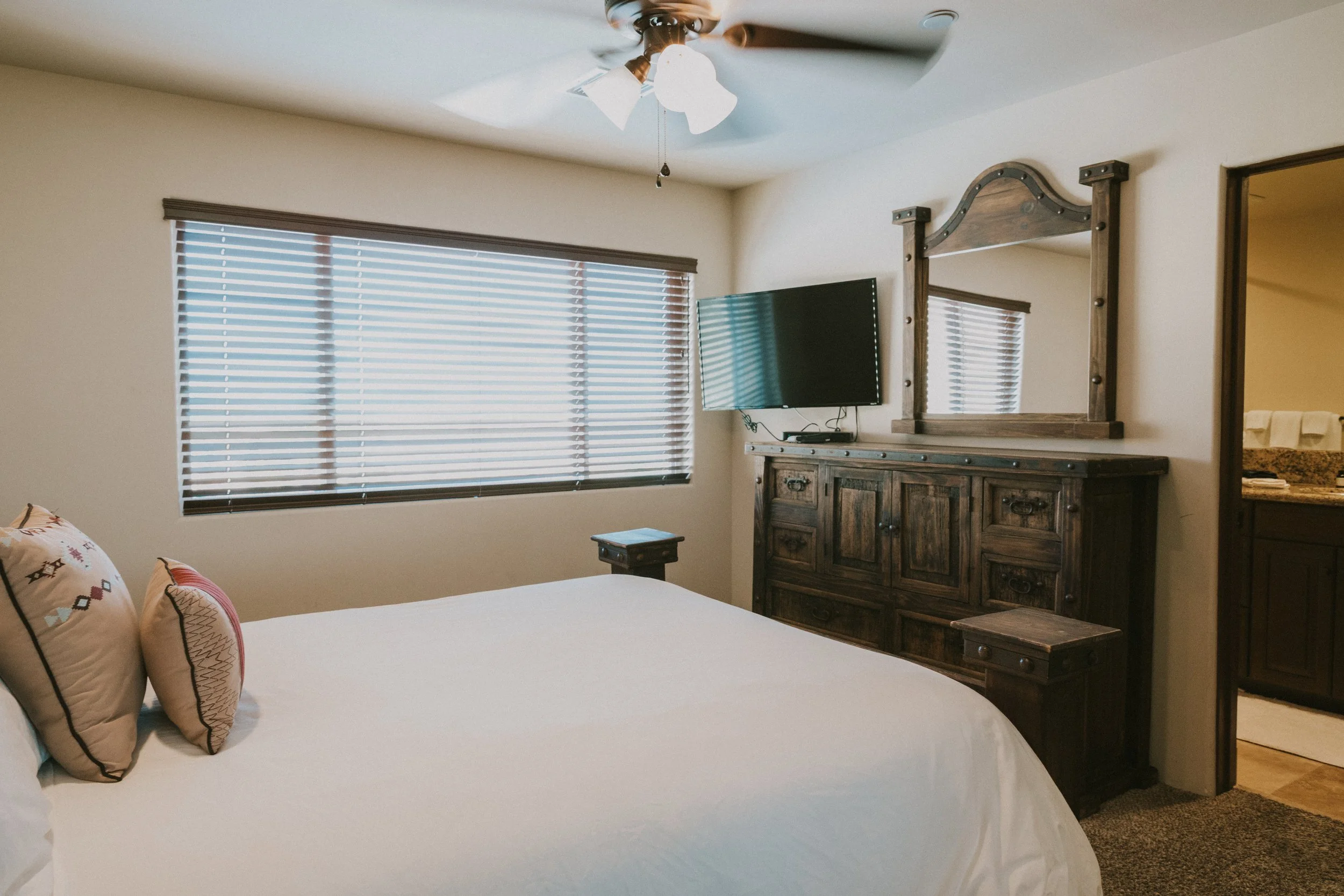 A bedroom with a bed, pillows, a wooden dresser with a mirror, a mounted TV, a window with blinds, and a ceiling fan with lights.
