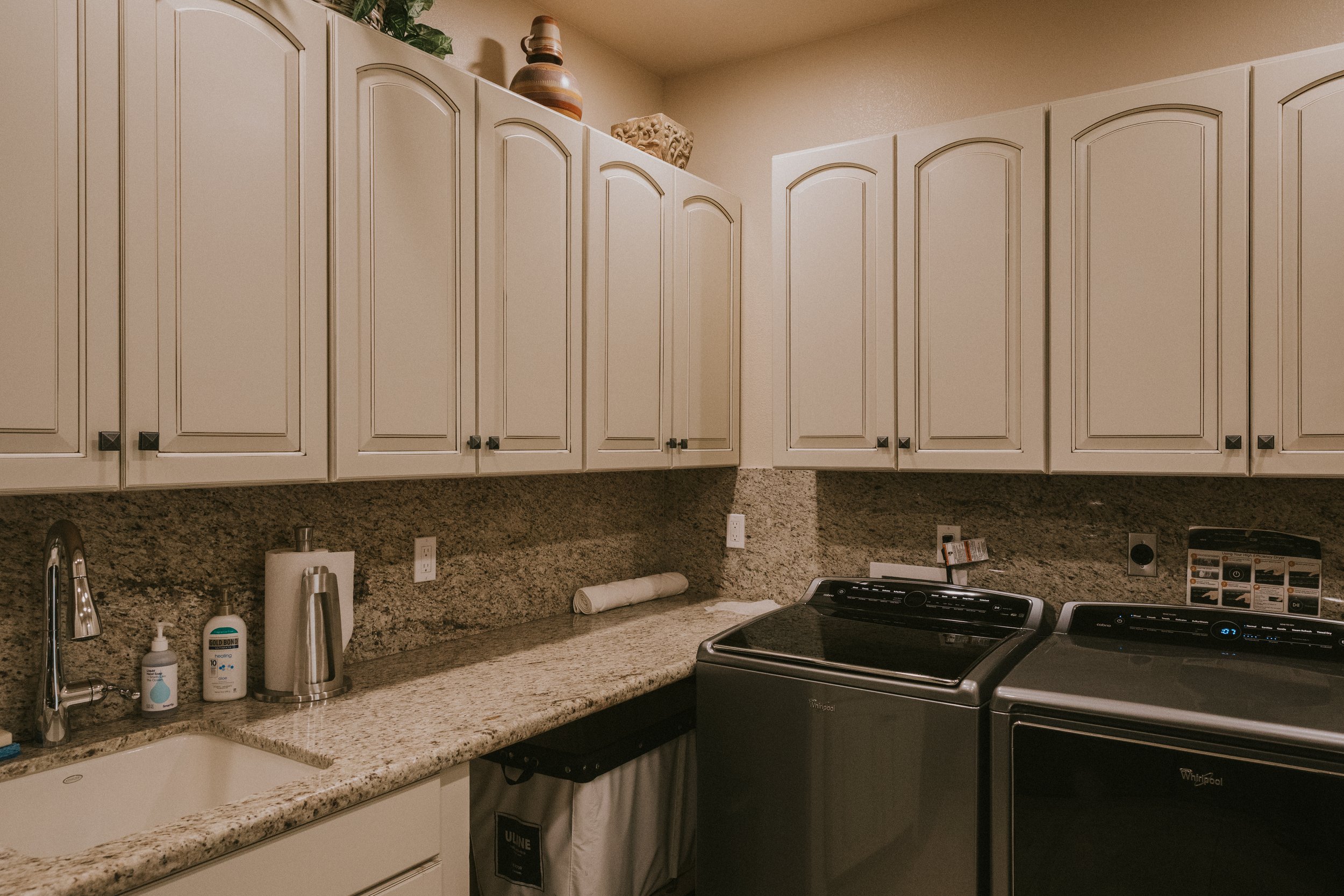 A kitchen with white upper cabinets, a granite countertop, a stainless steel sink, a soap dispenser, a hand sanitizer, a paper towel roll, a soap bottle, and a modern electric stove. Decorative vases and a stone sculpture are on top of the cabinets.