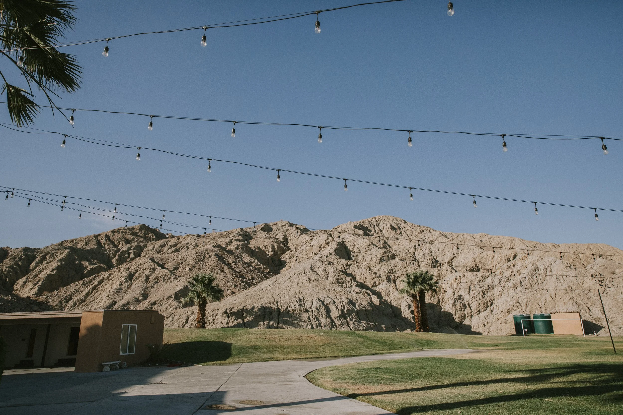 A mountain range with rugged, tan-colored peaks under a clear blue sky, with strings of outdoor lights hanging overhead, a palm tree on the left, and a small building with a curved sidewalk and a lawn in the foreground.