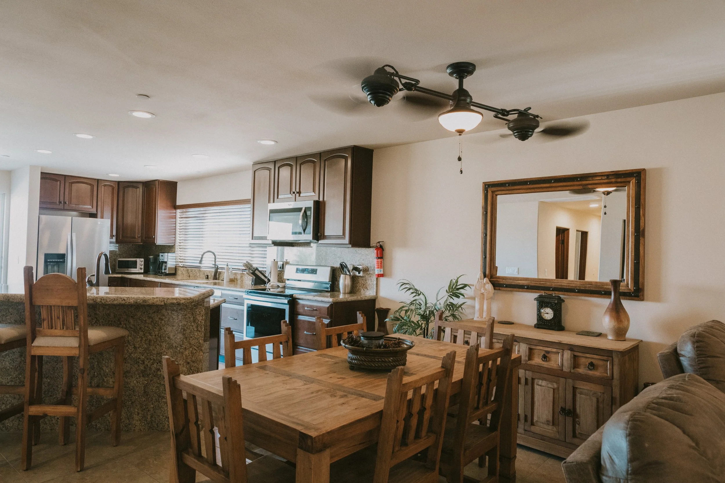 Open-concept kitchen and dining area with wooden furniture, granite countertops, stainless steel appliances, a large mirror, and a ceiling fan.