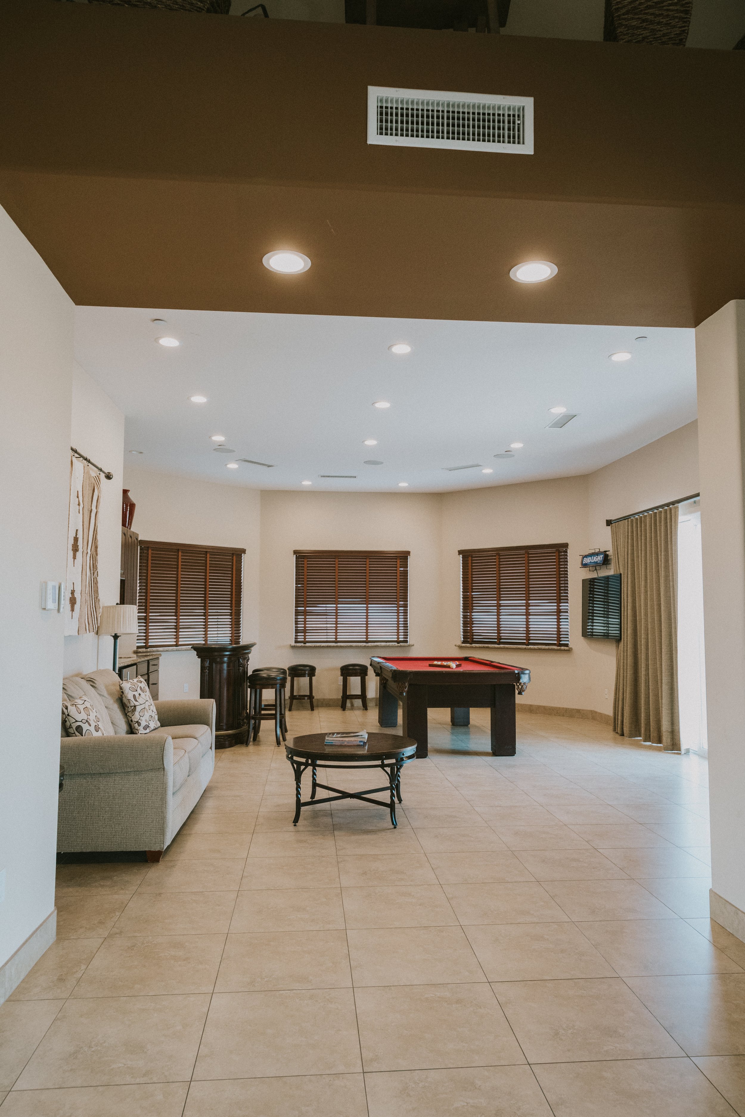 Living room with beige tile flooring, a beige sofa with patterned cushions, a black table with magazines, several windows with wooden blinds, a pool table, a wall-mounted TV, and curtains.
