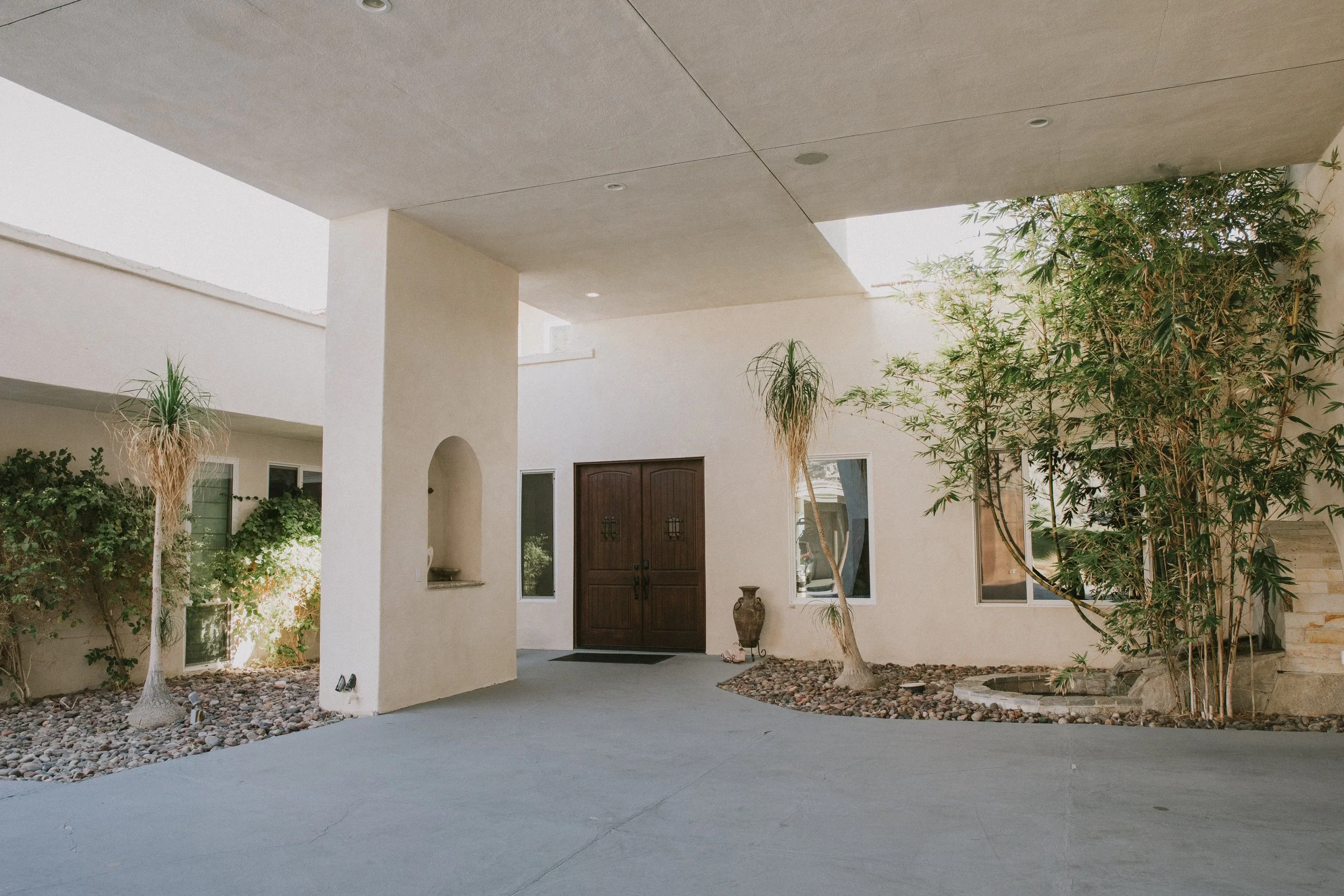 Front porch of a modern house with a flat roof, large wooden double door, and a small decorative vase near the door. There are trees with greenery and rocks lining the pathway, and a concrete driveway in the foreground.