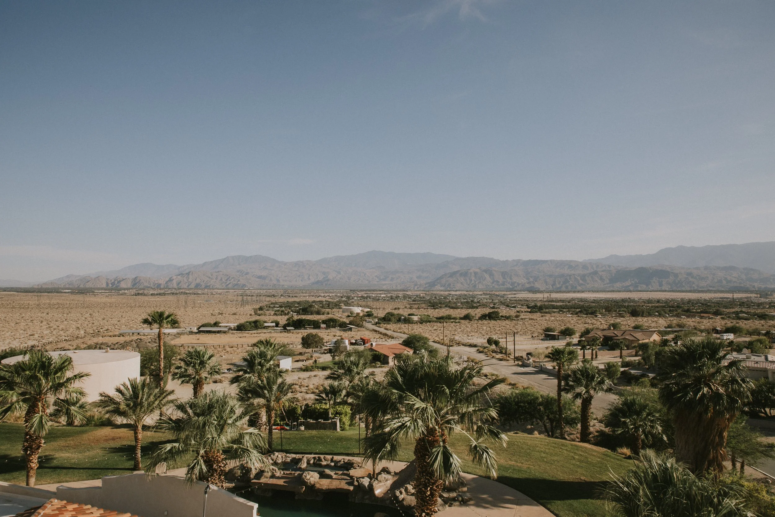 A desert landscape with mountains in the background and a residential area with palm trees and houses in the foreground.