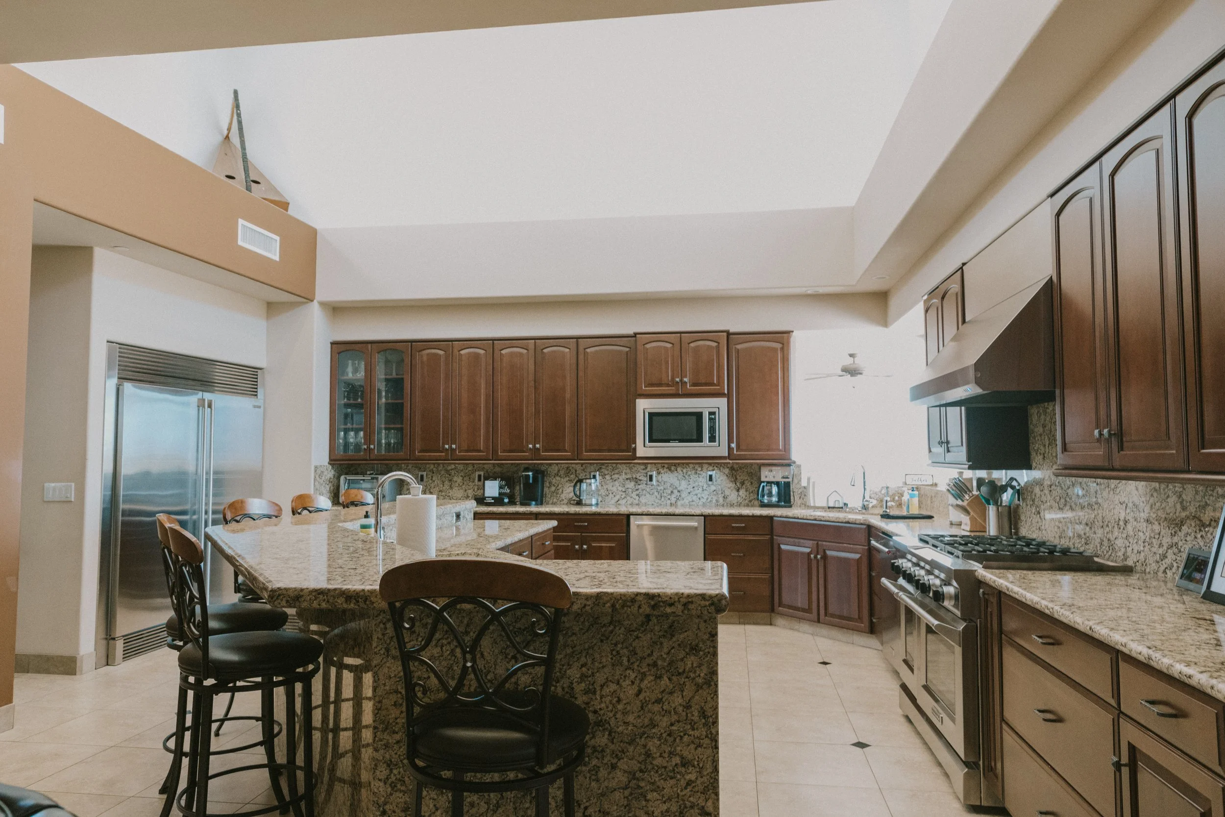 A spacious kitchen with wooden cabinets, granite countertops, a large kitchen island with chairs, stainless steel appliances, and a tiled floor.