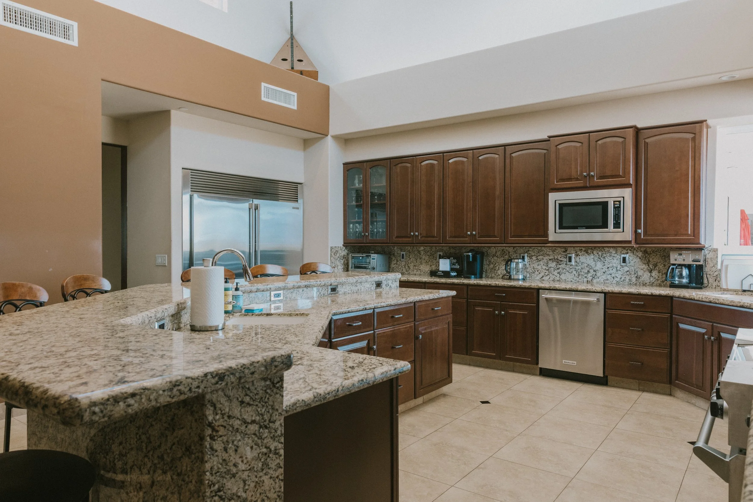 Modern kitchen with granite countertops, wooden cabinets, stainless steel appliances, and a large island with seating. There's a microwave, coffee maker, and various small appliances on the counters.