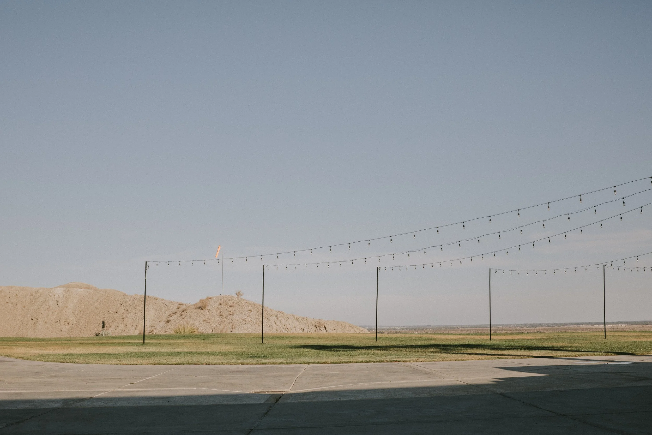 Outdoor scene with strung string lights hanging above a grassy area and a sandy hill, clear blue sky in the background.