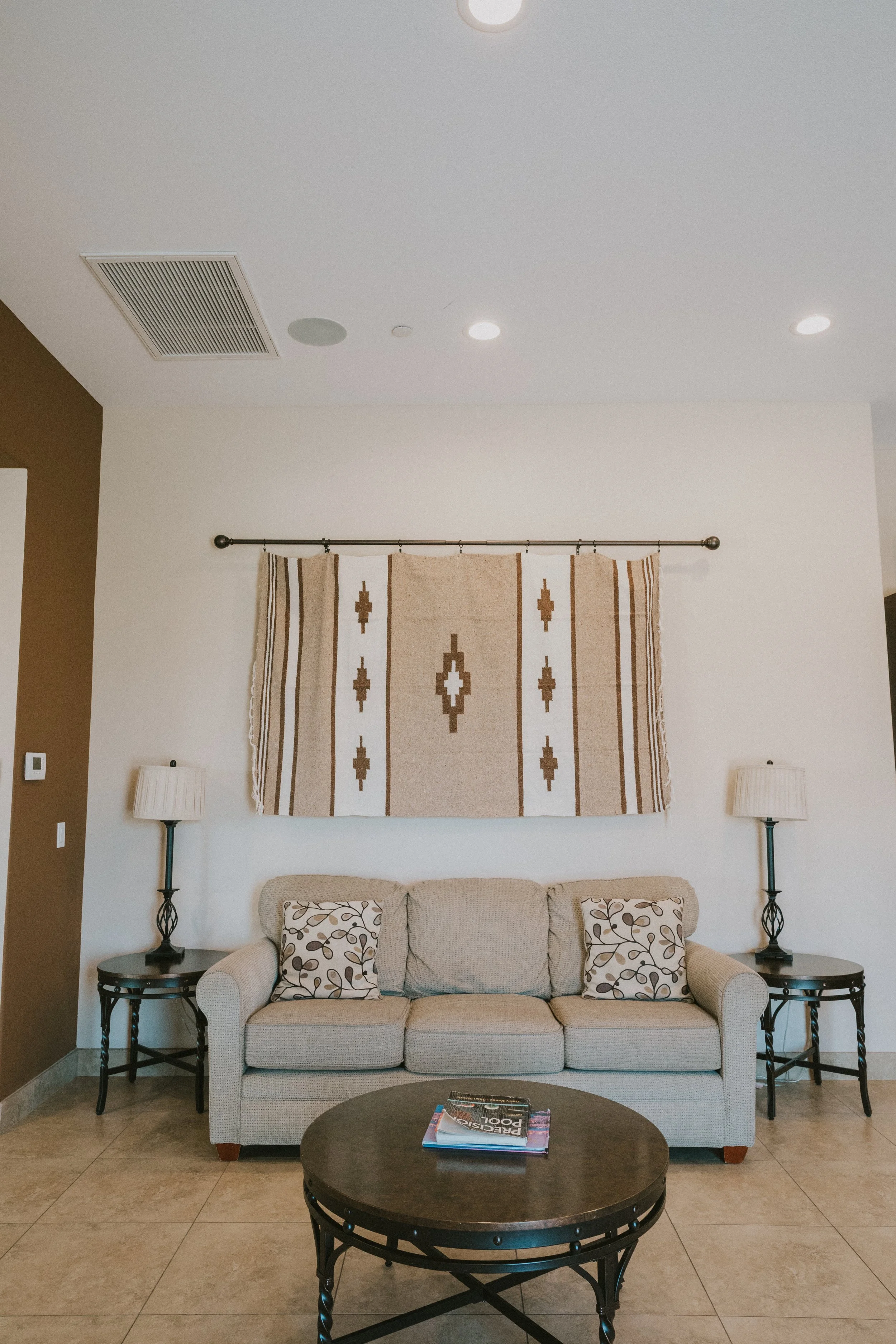 Living room with beige sofa, patterned pillows, matching table lamps, wall hanging with geometric design, and round coffee table with magazines.